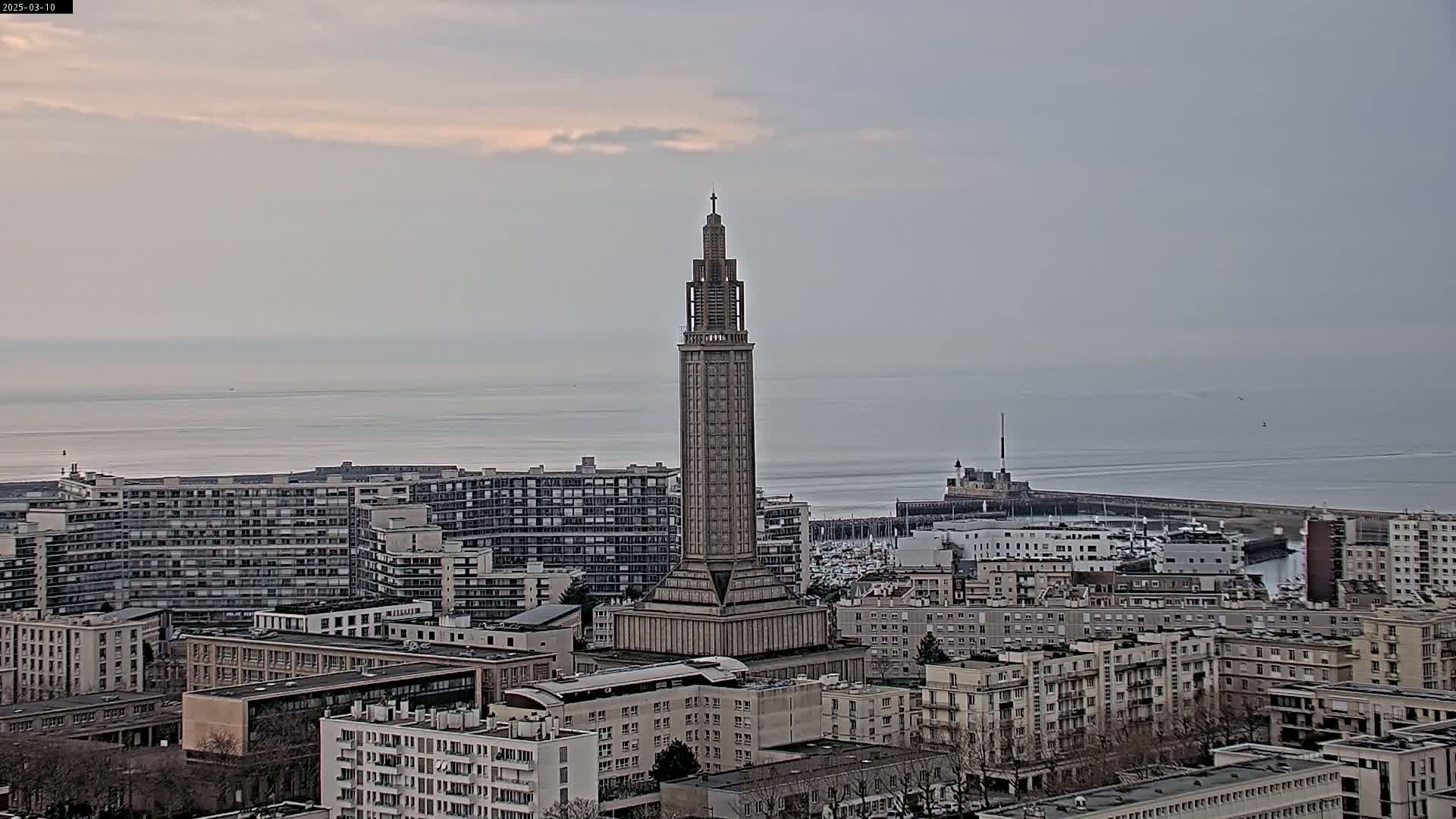 A tall, slender tower stands amidst a cityscape overlooking a calm sea under an overcast sky.