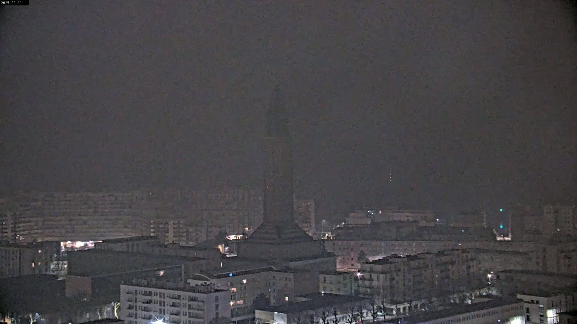 A tall, dark structure is visible in the distance amongst a city shrouded in heavy fog at night.