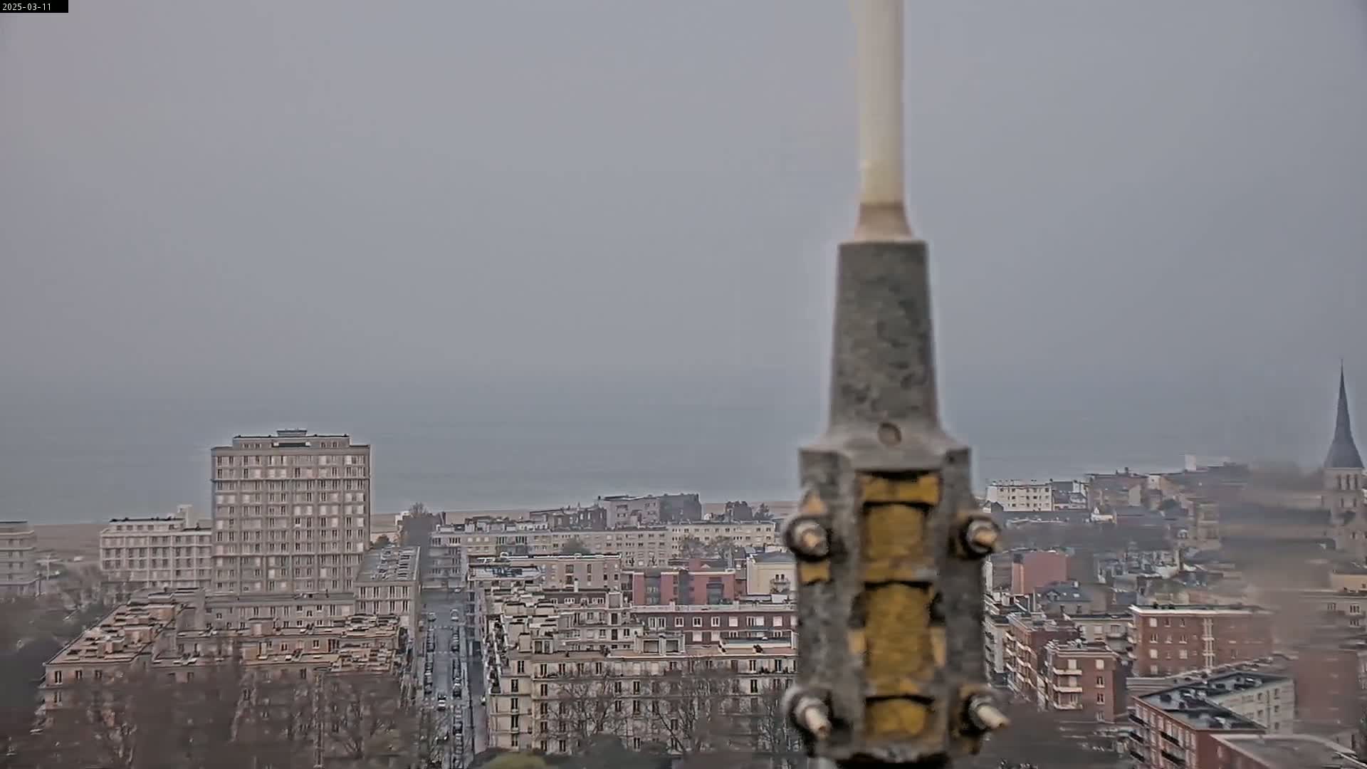 A hazy, overcast view of a city by the sea, seen from behind a tall, metallic structure.