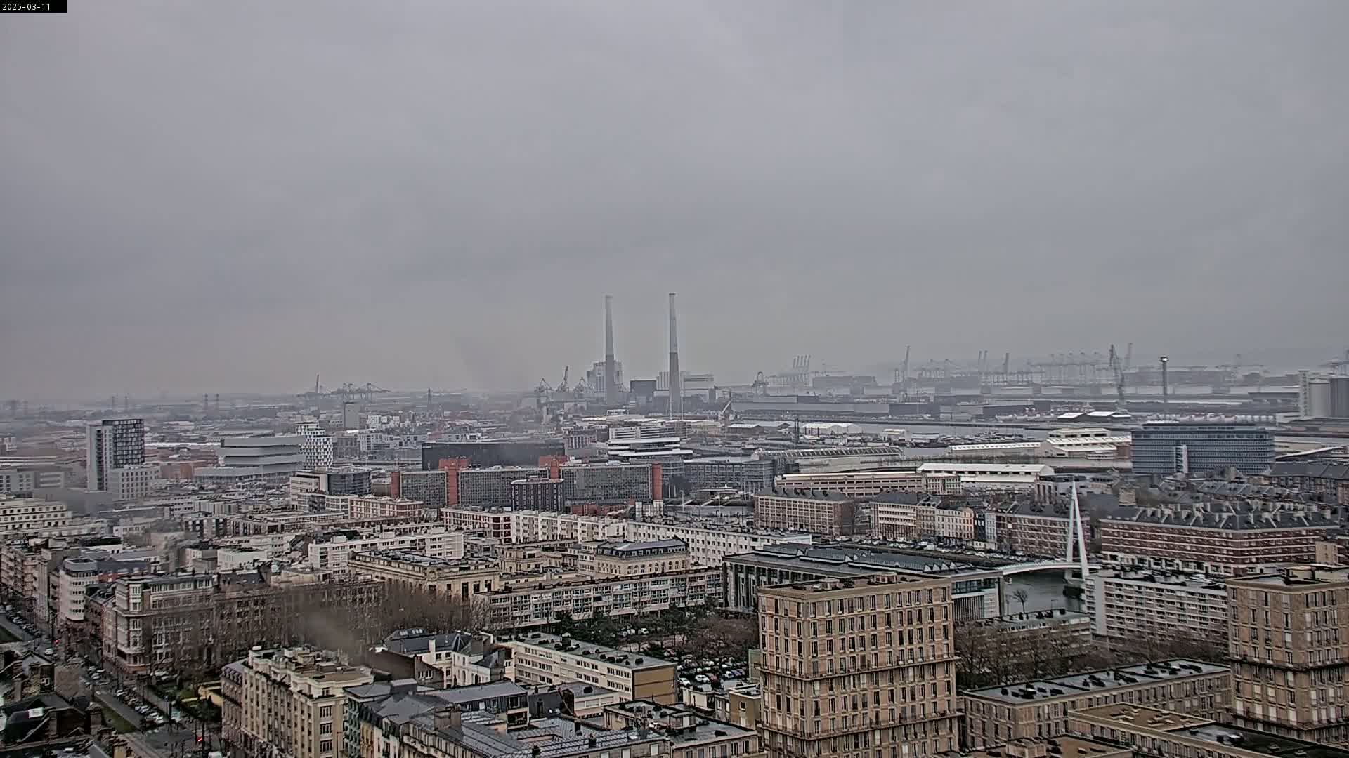 A hazy, overcast day reveals a sprawling cityscape with industrial areas and a port in the distance, all viewed from a high vantage point.