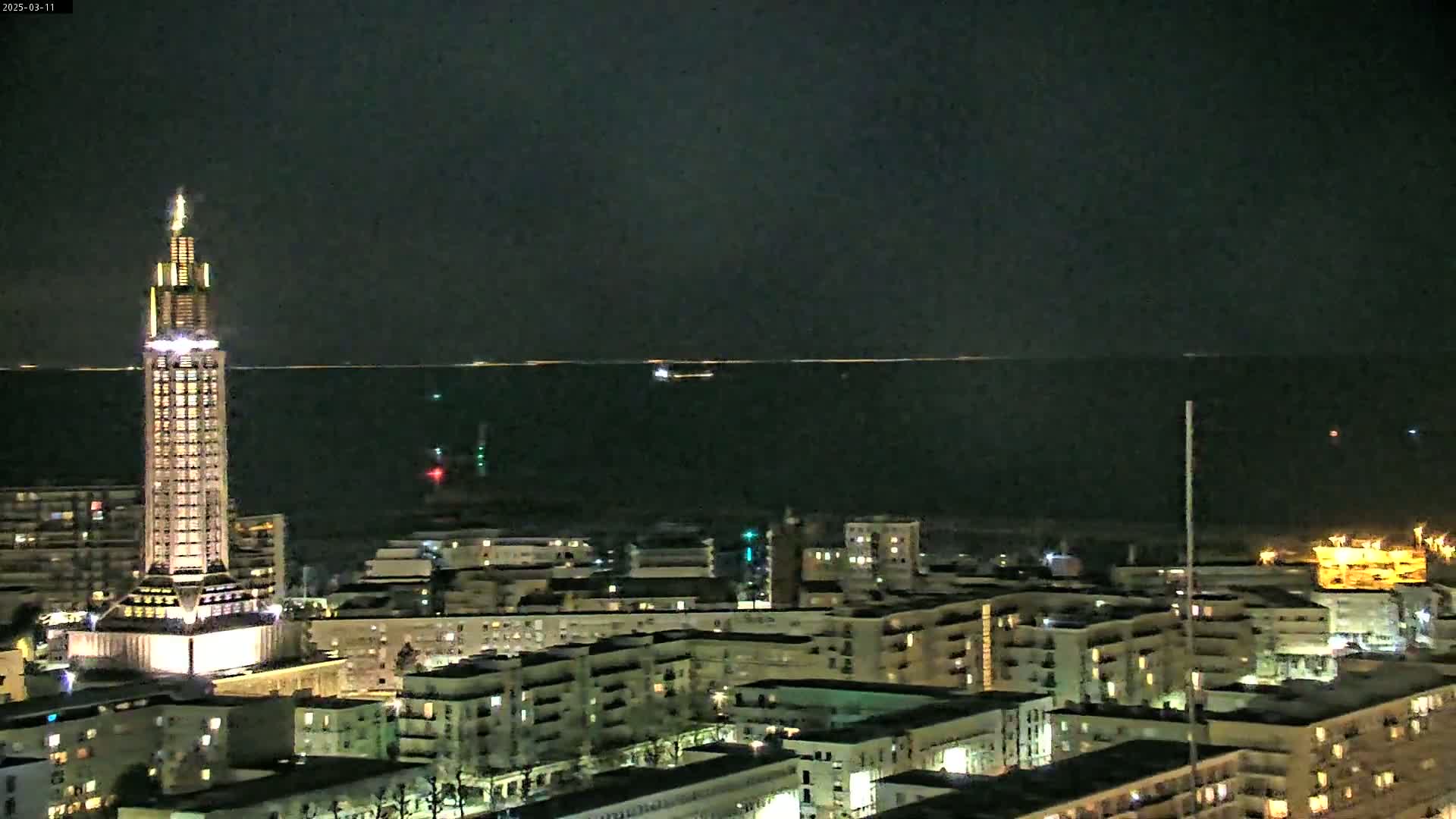 A brightly lit, tall tower stands prominently in the foreground of a nighttime cityscape overlooking a dark body of water under a clear, dark sky.