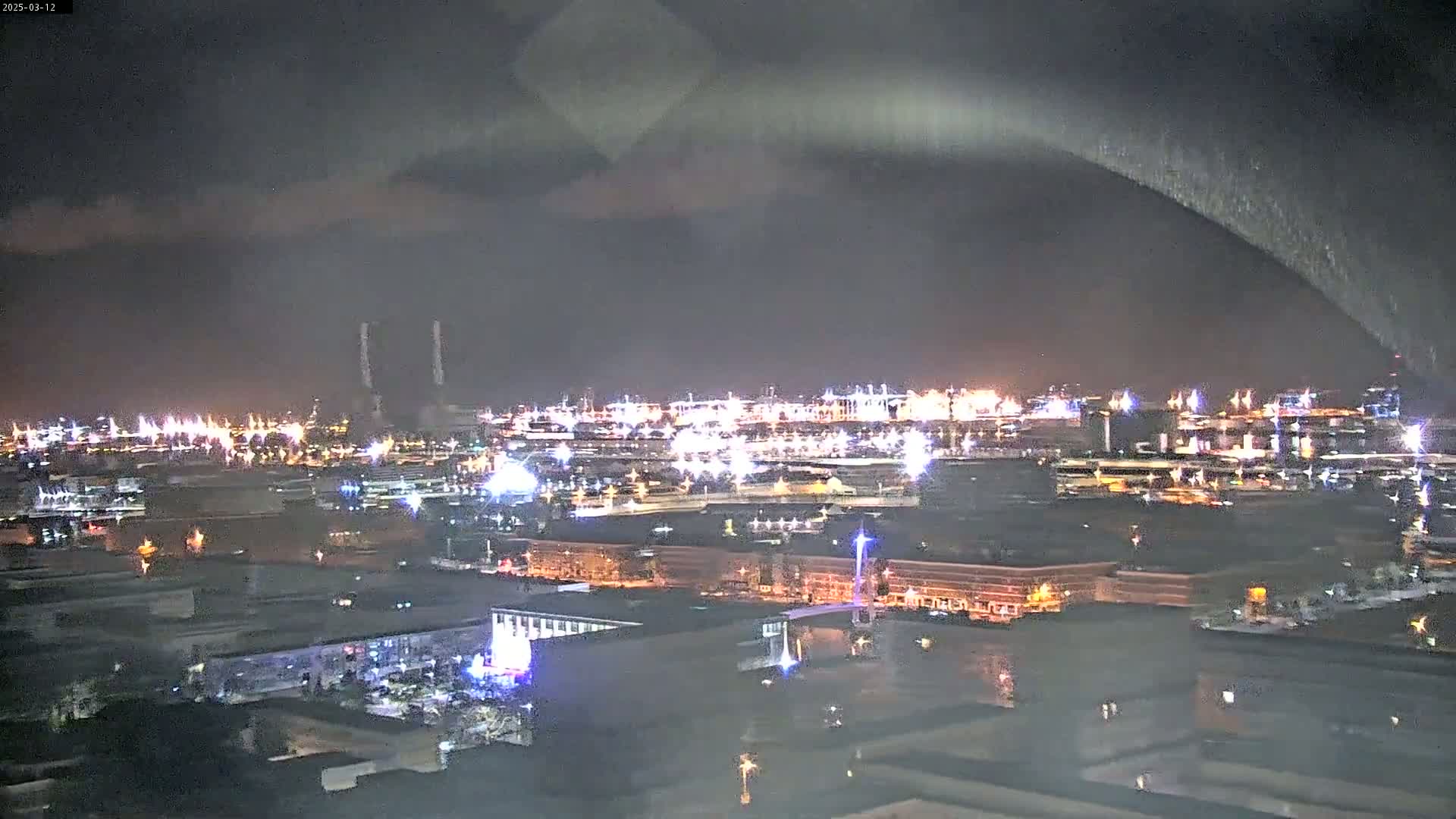 A nighttime view of an industrial area and city, illuminated by numerous lights under a dark, cloudy sky.
