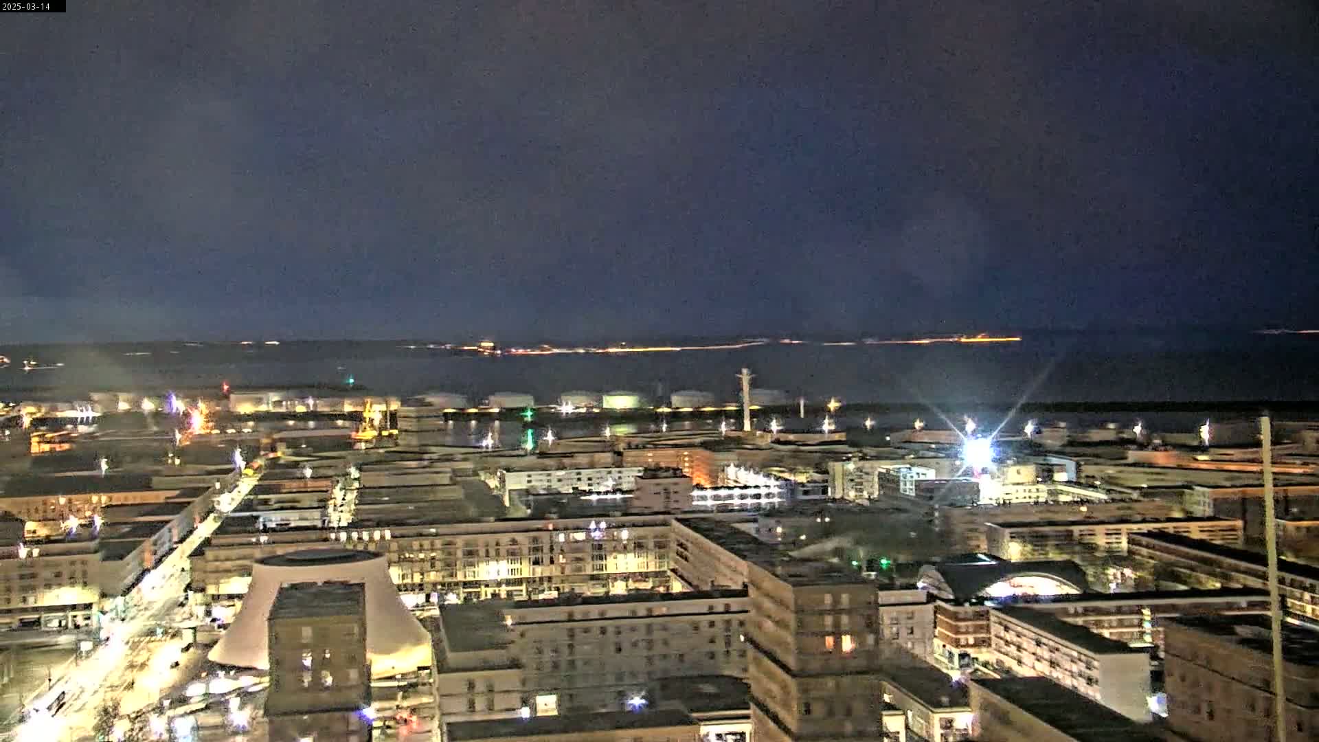 A nighttime aerial view of a city's harbor and waterfront area, with a dark, clear sky above and numerous lights illuminating the buildings and industrial structures.