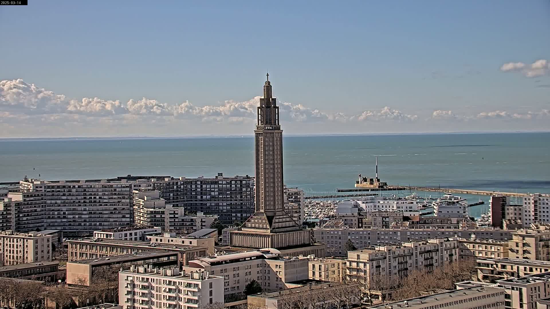 A tall, slender tower stands in a city overlooking a calm sea under a partly cloudy sky.