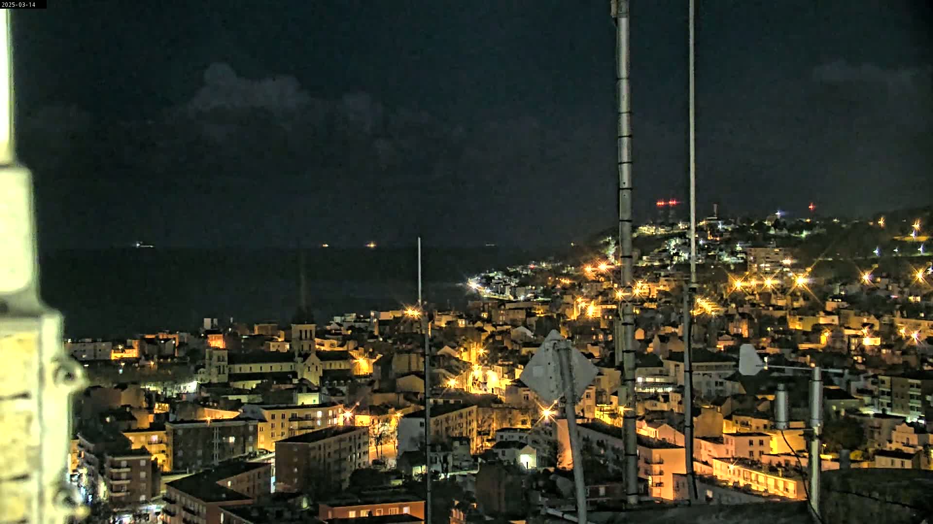 A nighttime view of a coastal city with its lights illuminated against a dark, cloudy sky and a calm sea.