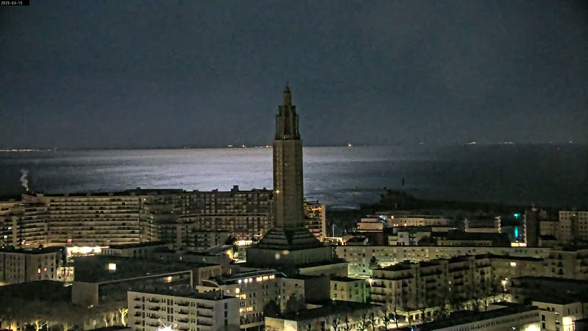A tall tower stands in a city at night overlooking a calm, dark body of water under a dark sky.