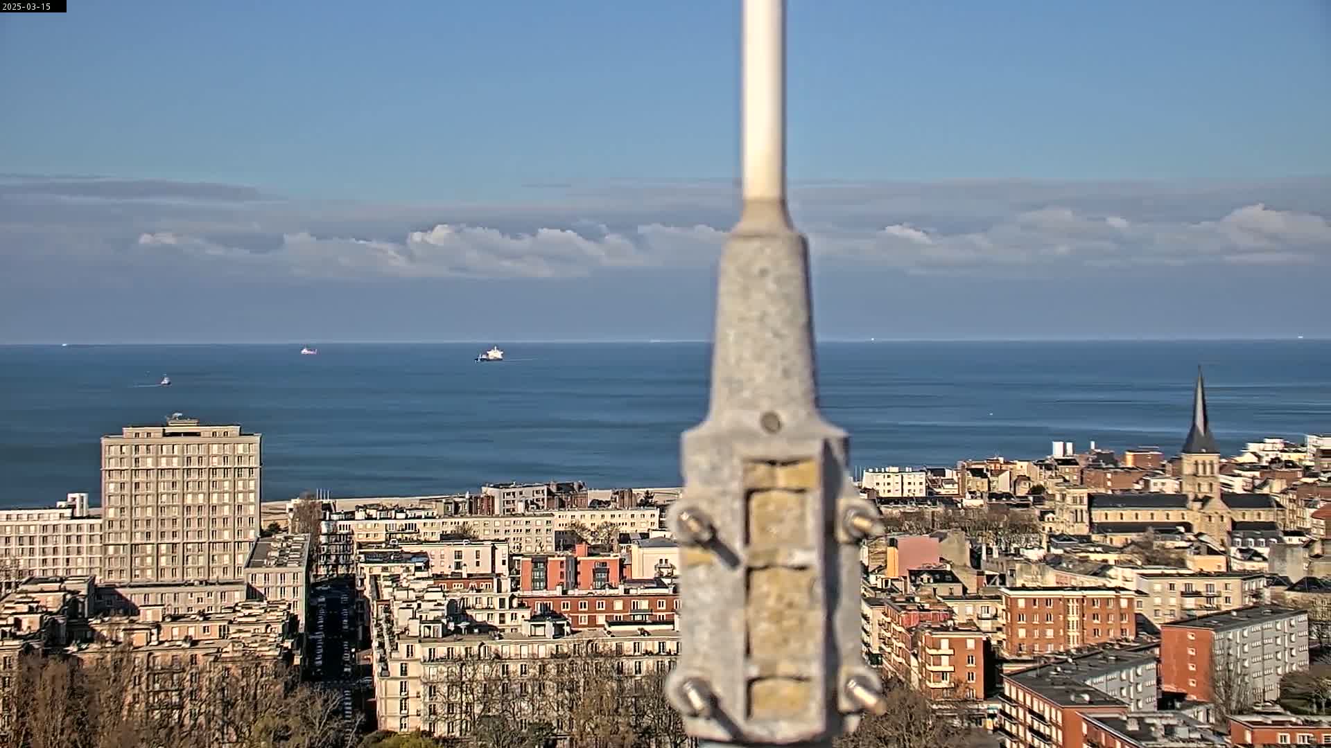 A mostly clear sky overlooks a coastal city with a calm ocean in the background, a tall metal post centered in the foreground.