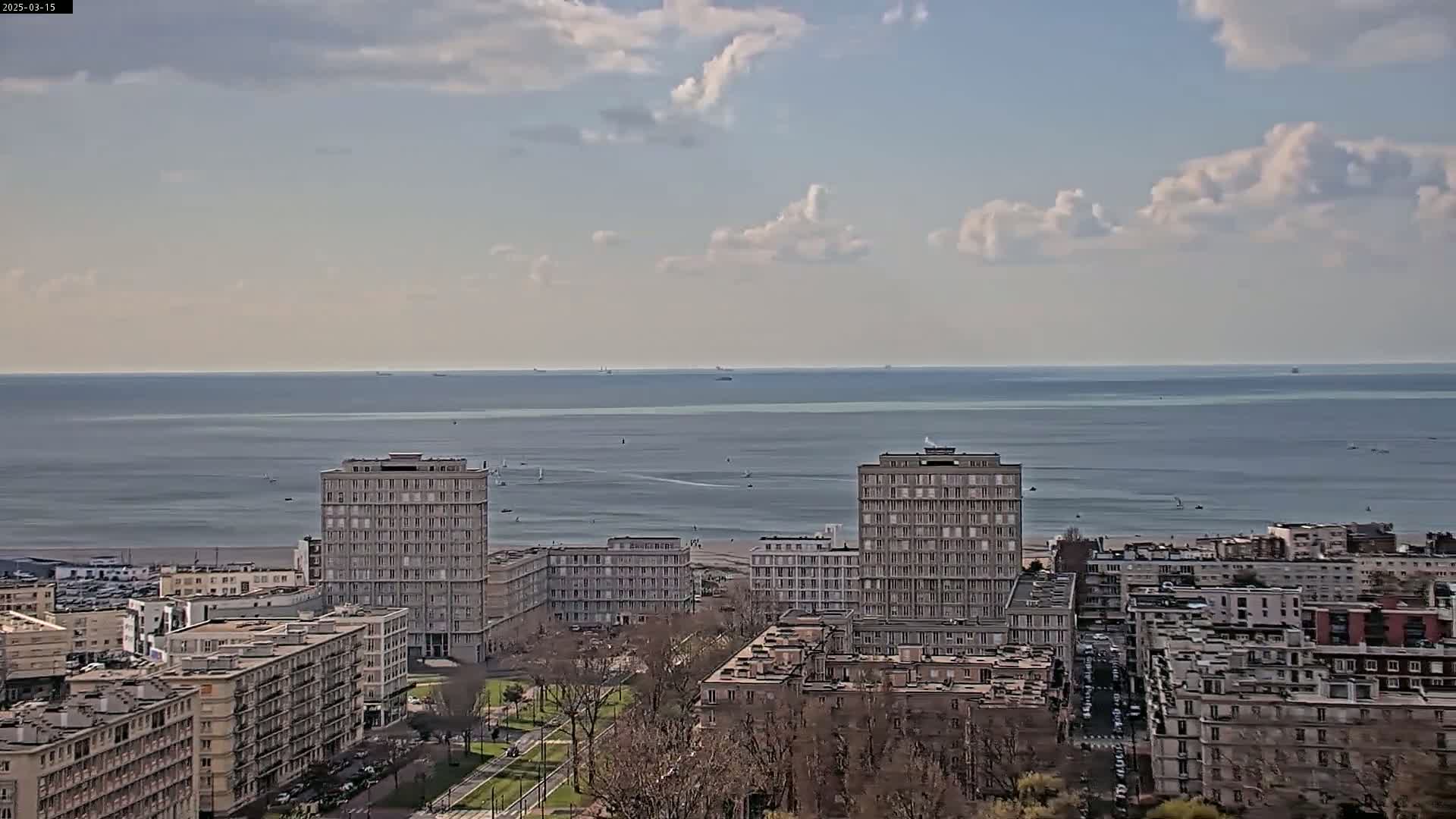 A coastal city scene under a partly cloudy sky shows several tall apartment buildings overlooking a beach and a calm ocean with a few boats visible on the horizon.