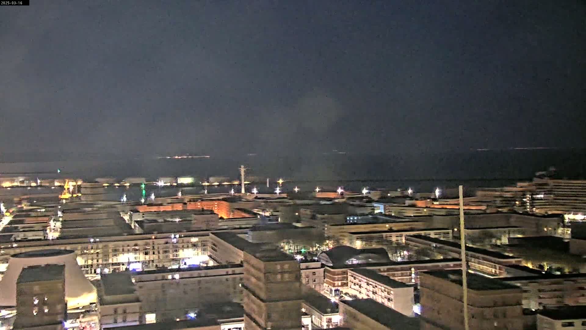 A nighttime, high-angle view of a city harbor with many lights illuminating buildings and the water, under a dark, clear sky.