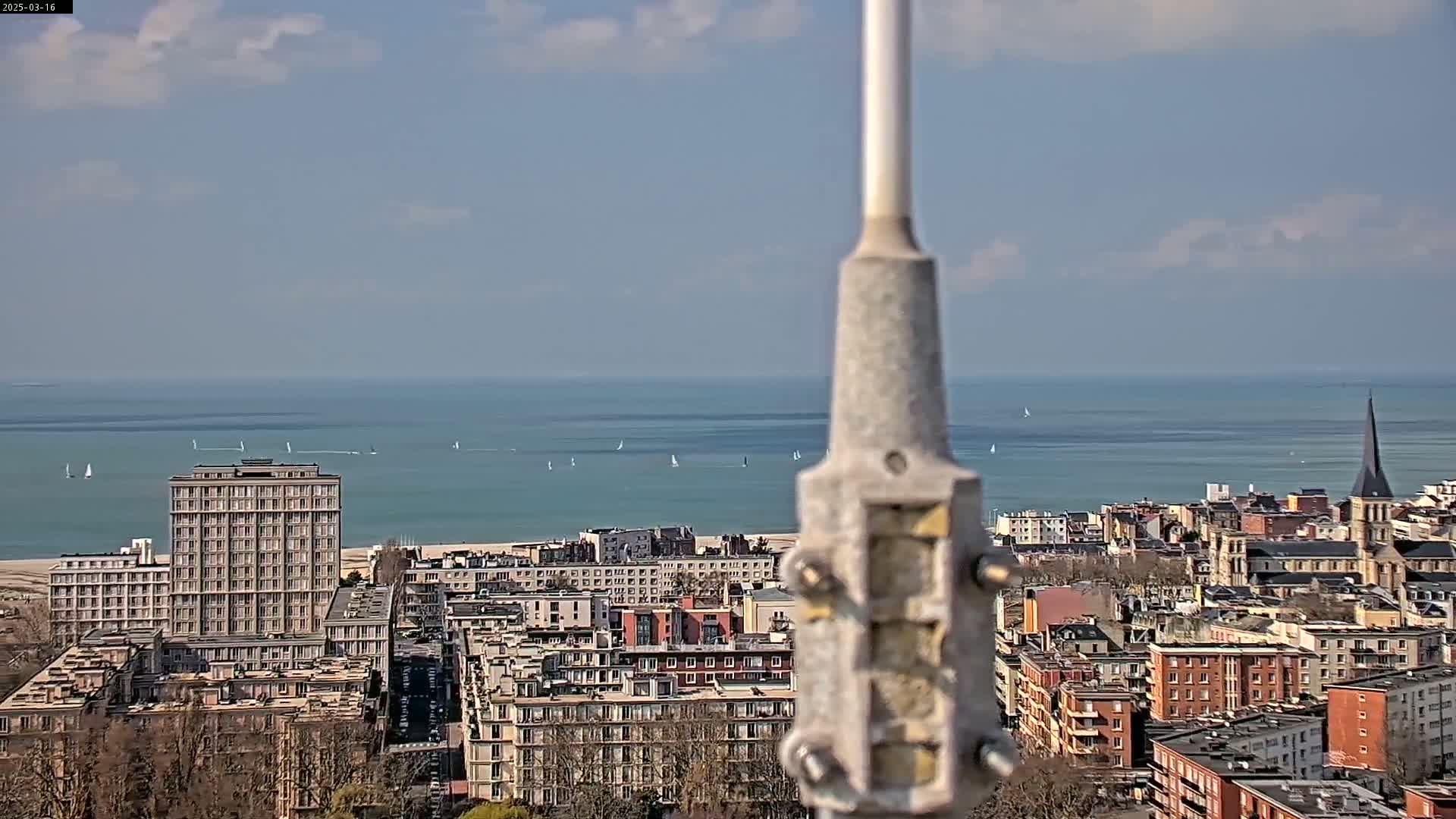 A partly cloudy day shows a coastal city with numerous buildings and several sailboats visible on the calm ocean in the distance, viewed from a high vantage point.