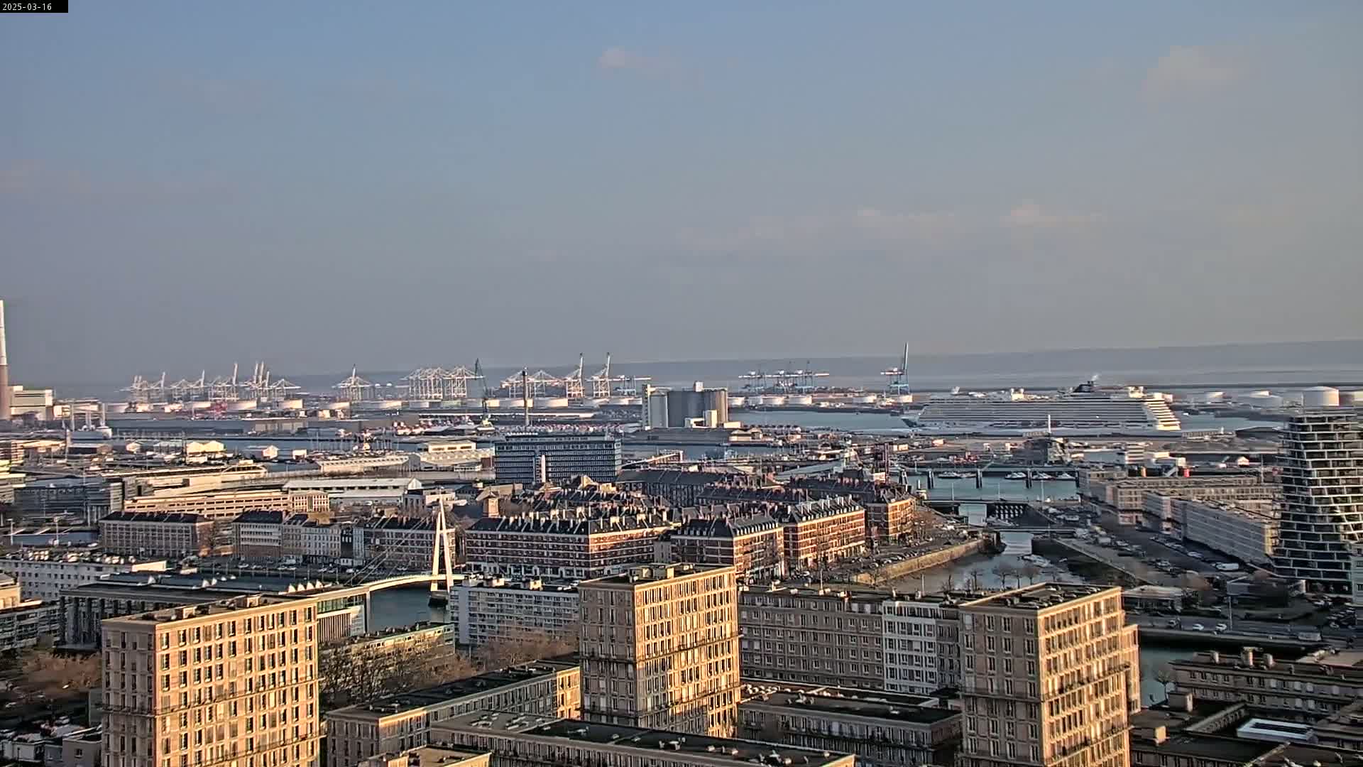 A cityscape with numerous buildings, a harbor filled with docked ships and cranes, and a cruise ship in the distance under a mostly clear sky.