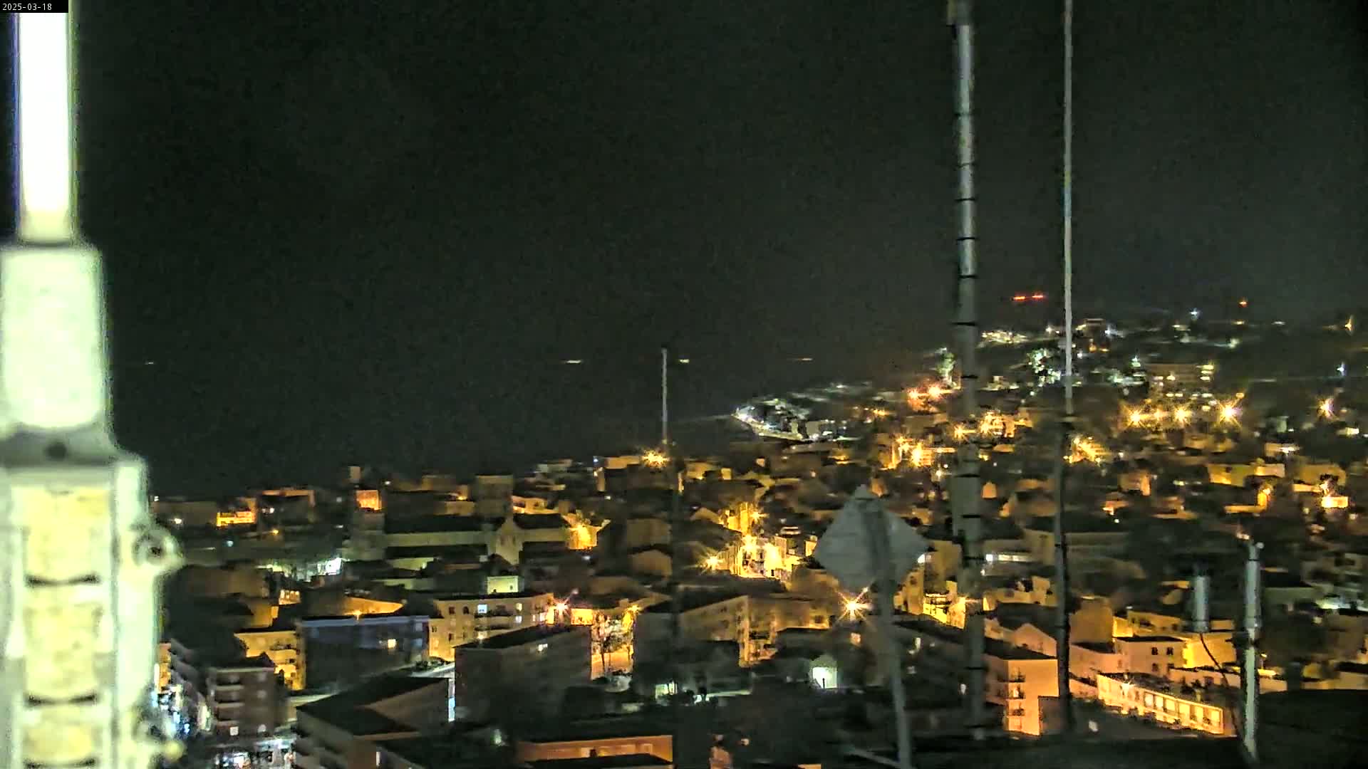 A nighttime, high-angle view shows a coastal town with numerous lights illuminating buildings and streets, extending to a dark body of water under a clear, dark sky.