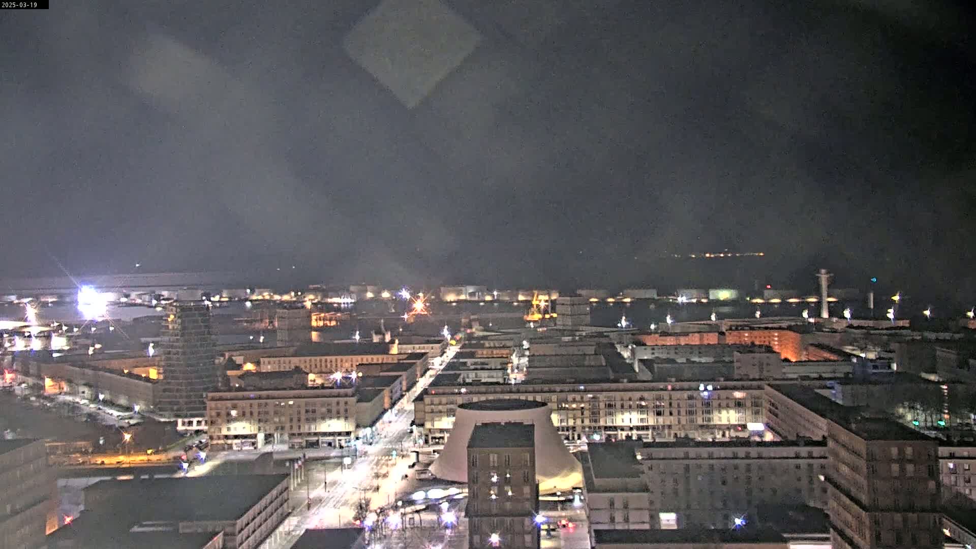 A nighttime aerial view of a city with a harbor in the background, under a dark, cloudy sky.