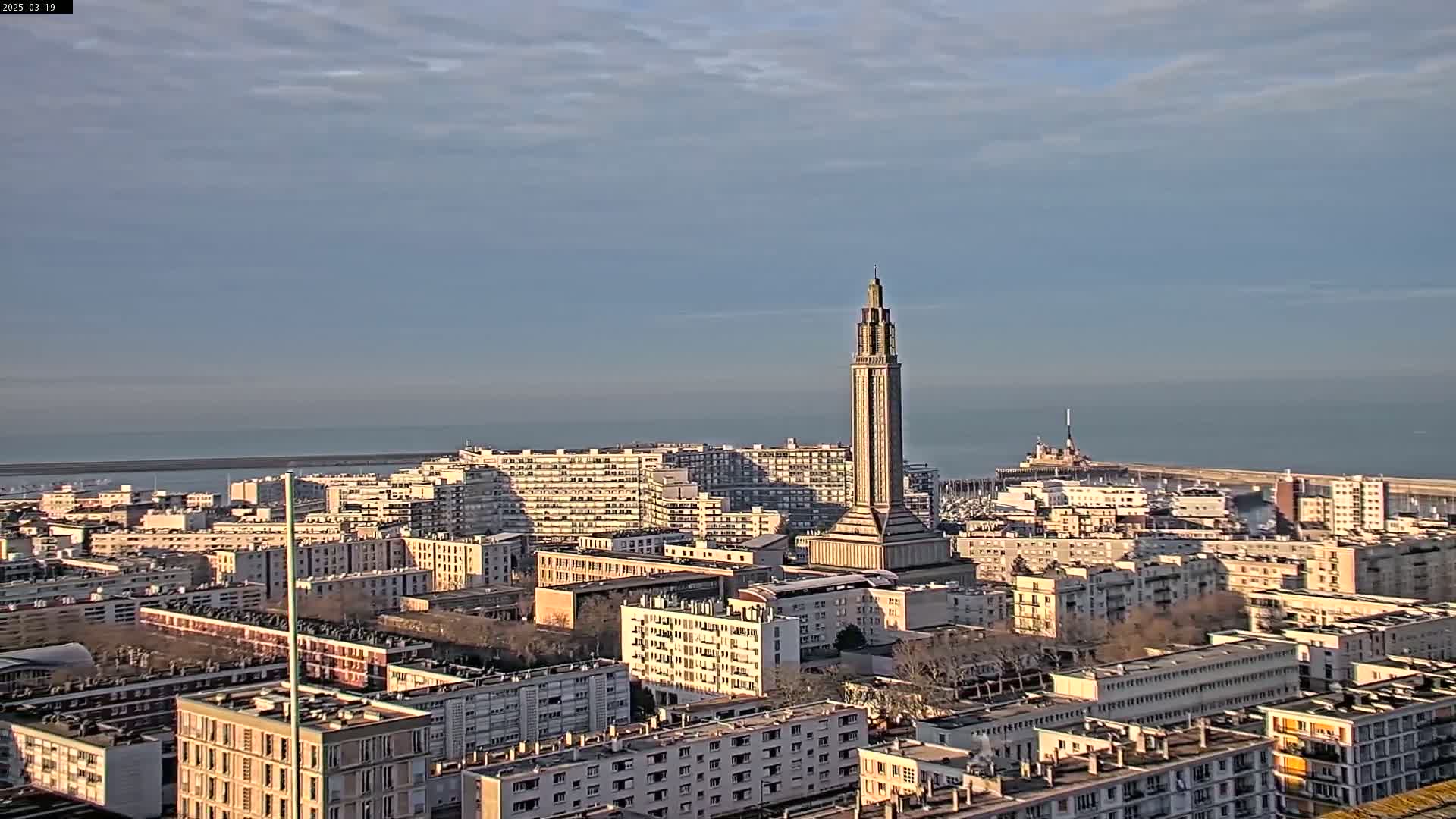 A city skyline with a tall, slender tower at its center is visible under a mostly clear, bright sky, overlooking a calm body of water in the distance.