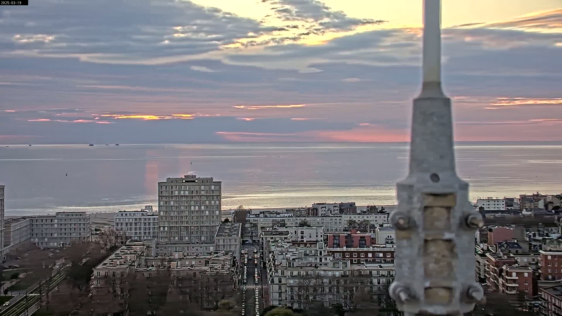 A coastal city at sunrise, with a calm sea and a partly cloudy sky, is viewed from an elevated position behind a tall metal post.