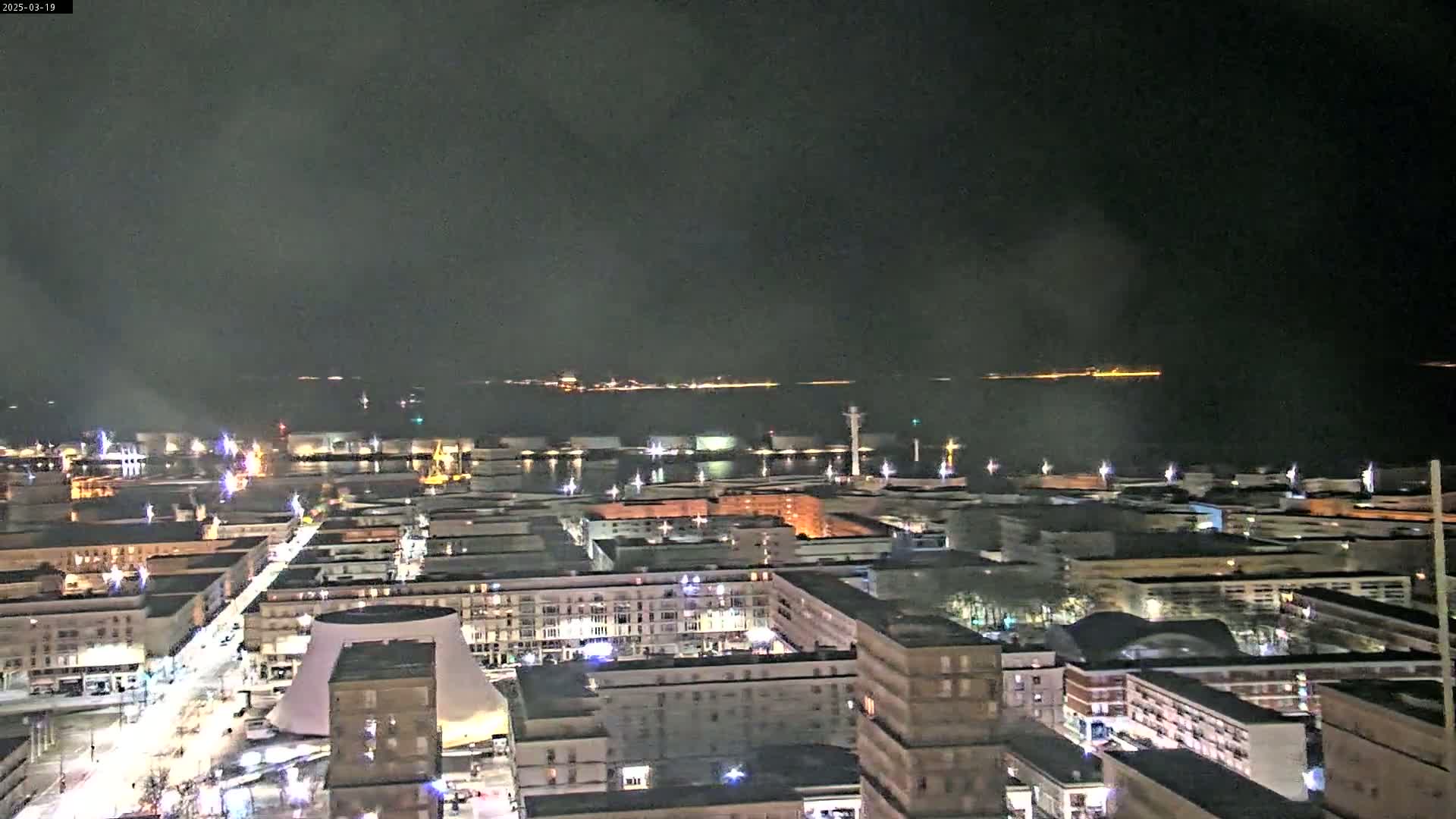 A nighttime, high-angle view shows a city harbor with its lights illuminated, and a dark body of water beyond it under a cloudy sky.