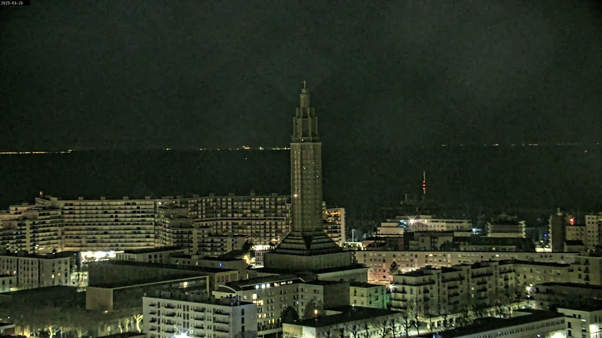 A tall, slender tower stands prominently in a nighttime cityscape overlooking a dark body of water under a clear, dark sky.