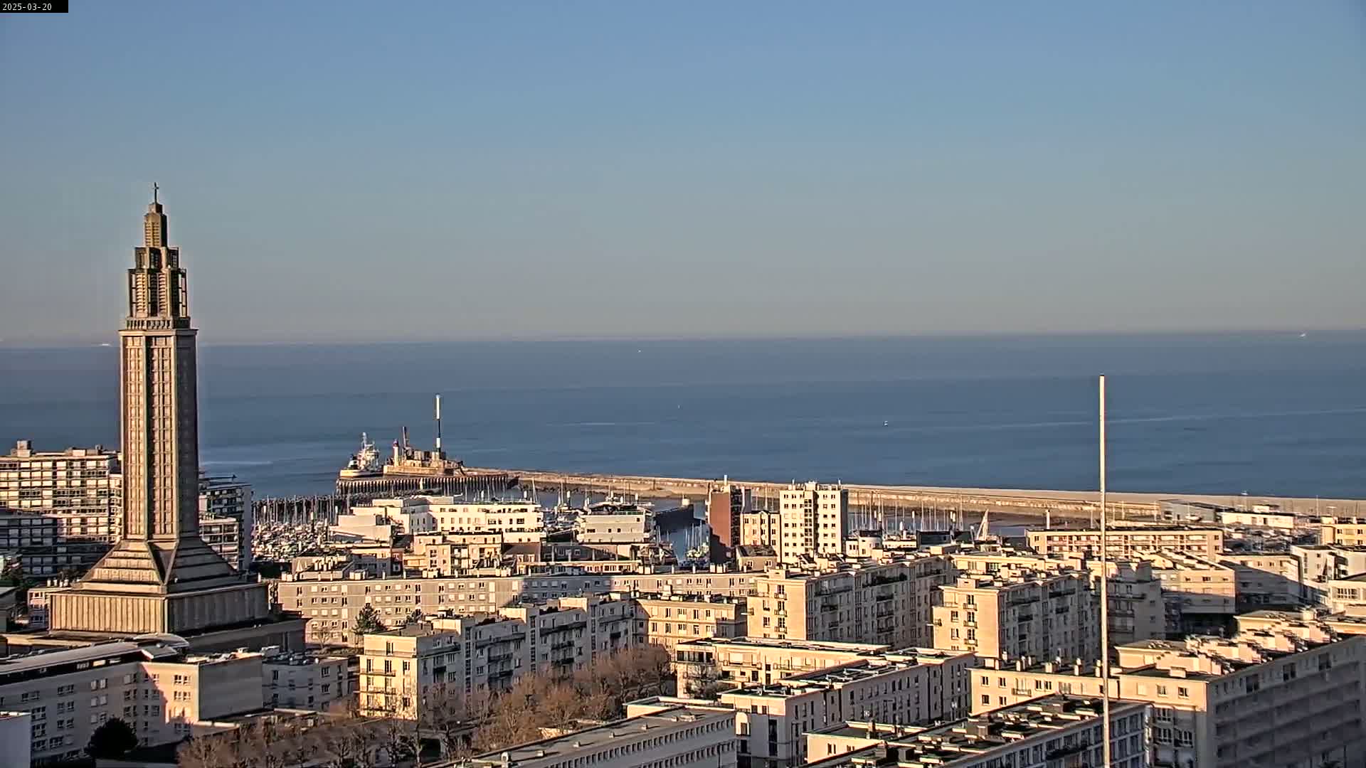 A tall, slender tower stands in the foreground overlooking a city by a calm ocean on a clear day.