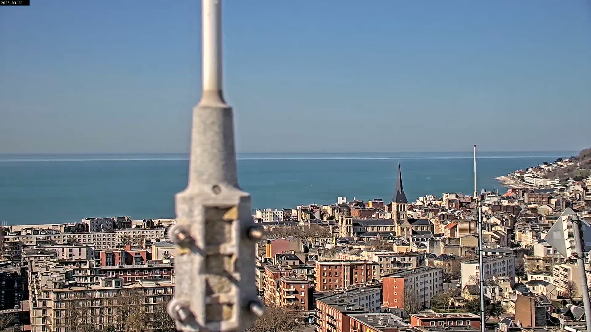 A coastal city with a church steeple is visible in the distance, under a clear blue sky, with a calm blue sea beyond it.