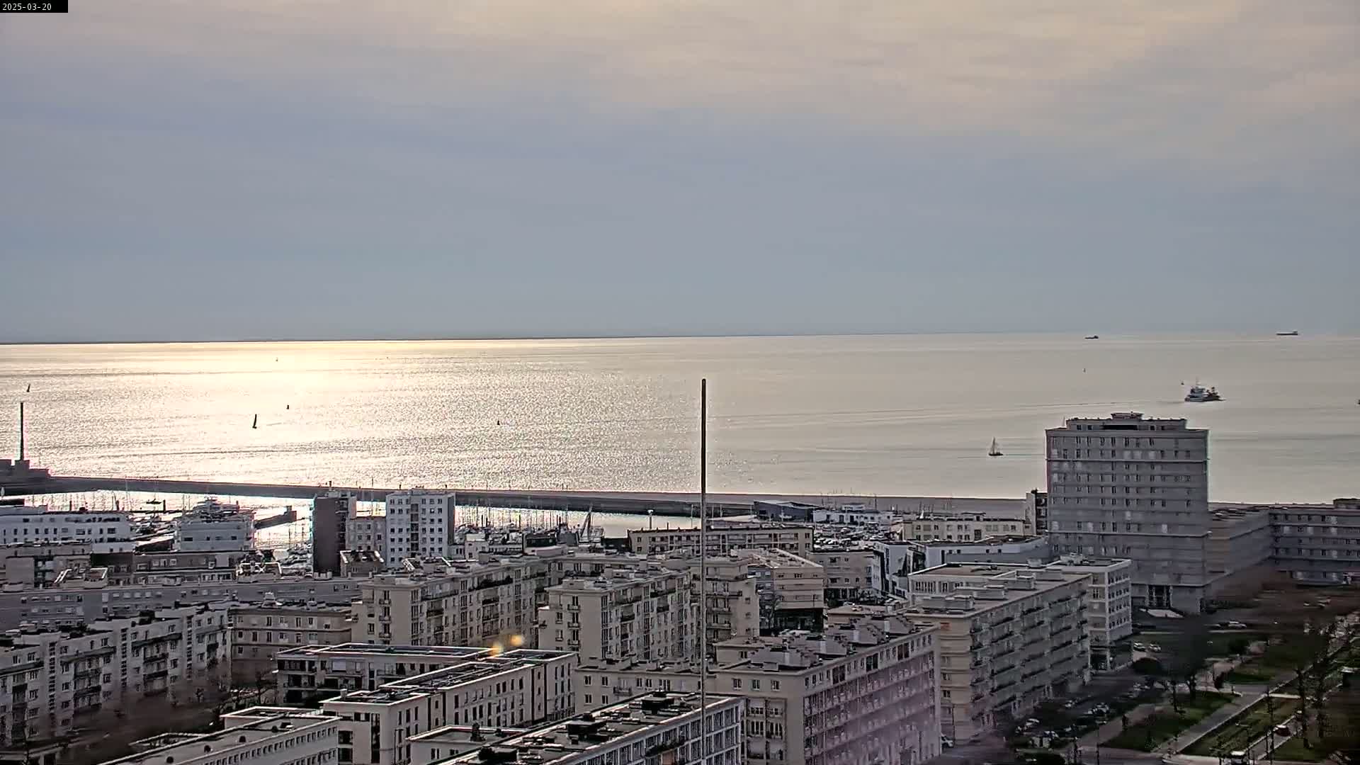 A glittering expanse of sea under a mostly cloudy sky is viewed from above a city of closely-packed apartment buildings near a harbor.