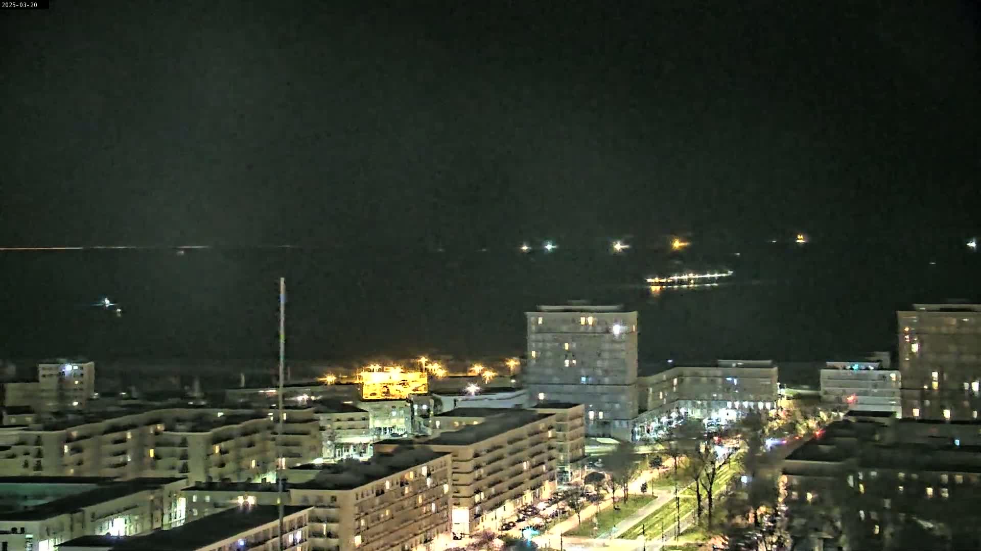 A nighttime view of a city waterfront shows multi-story buildings with illuminated windows overlooking a calm, dark body of water where several small lights are visible in the distance.