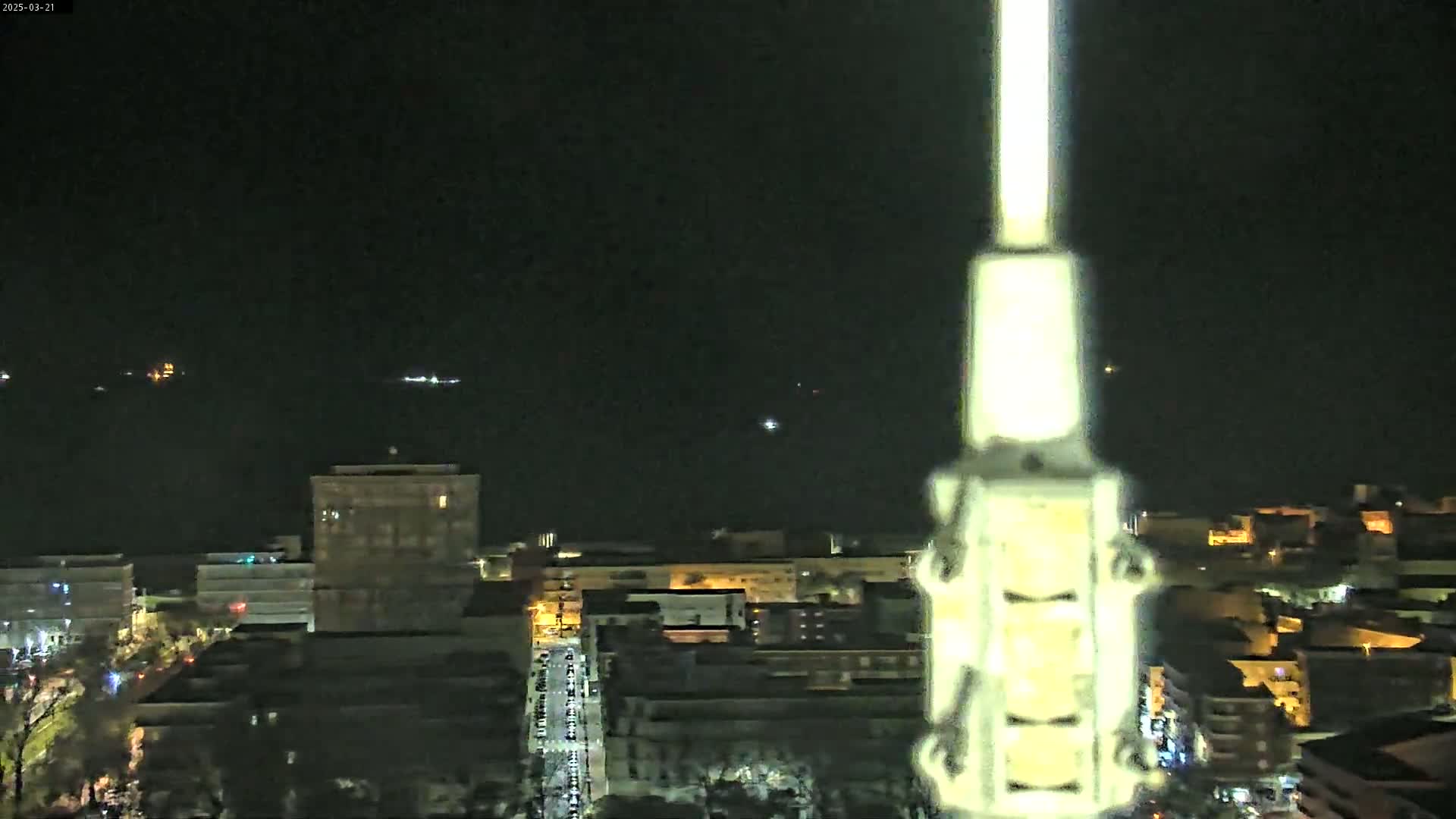 A nighttime view of a city skyline with dimly lit buildings and streets, and distant lights visible on the water under a dark sky.