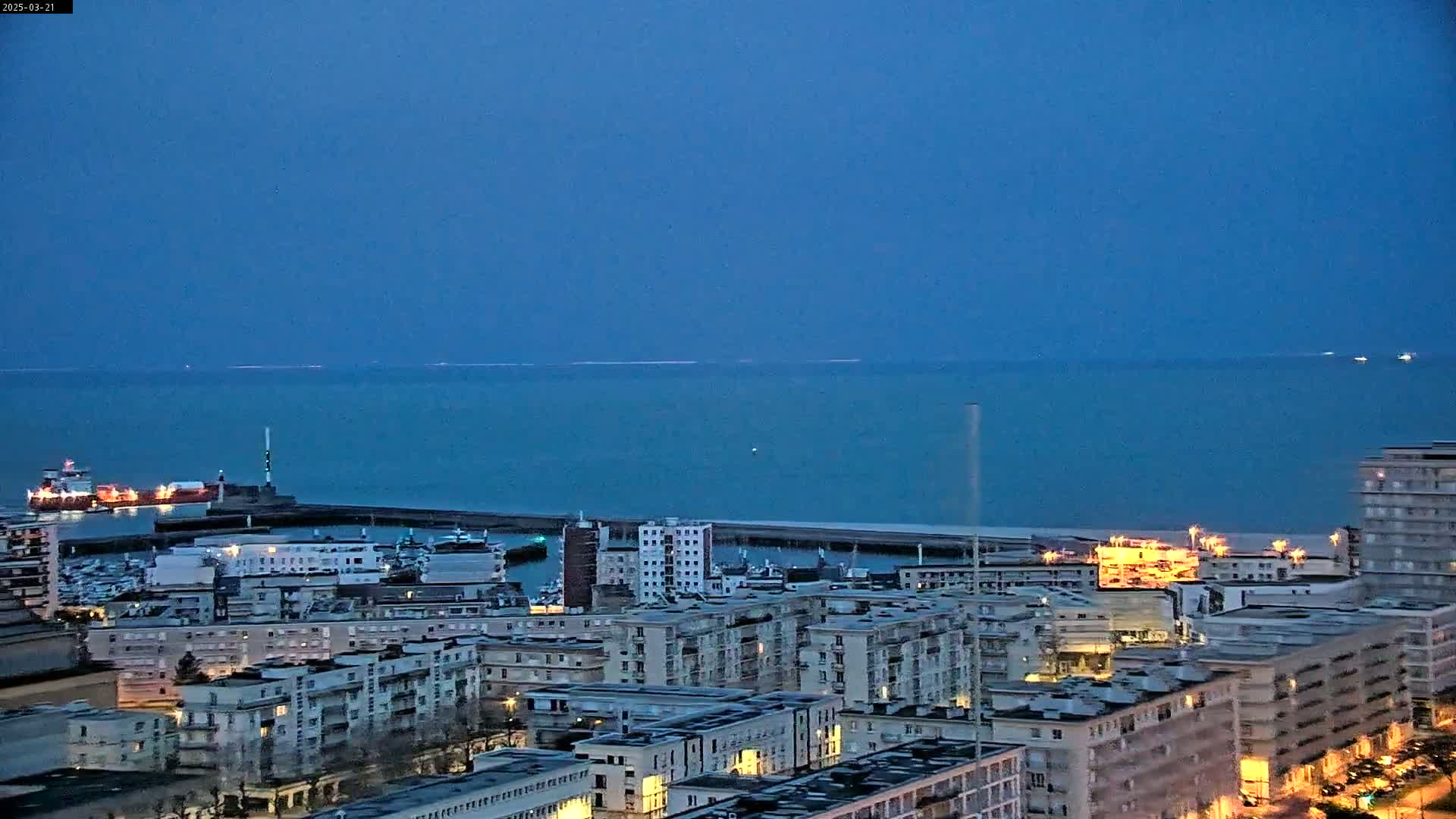 A city's waterfront at twilight, under a clear, dark blue sky, shows a harbor with ships and numerous buildings stretching into the distance, next to a calm body of water.