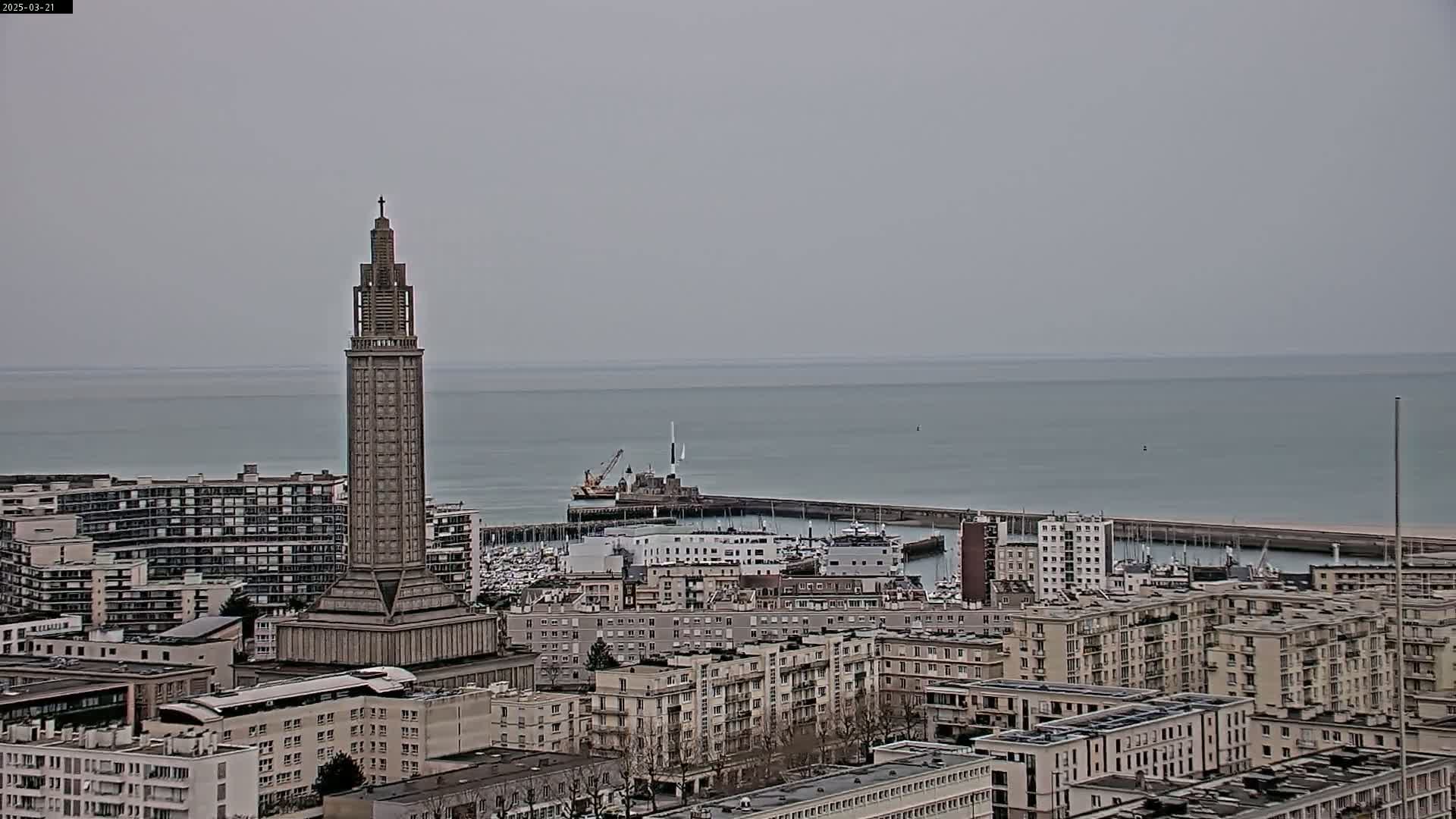 A tall, slender tower stands prominently in a cityscape overlooking a calm sea under an overcast sky.
