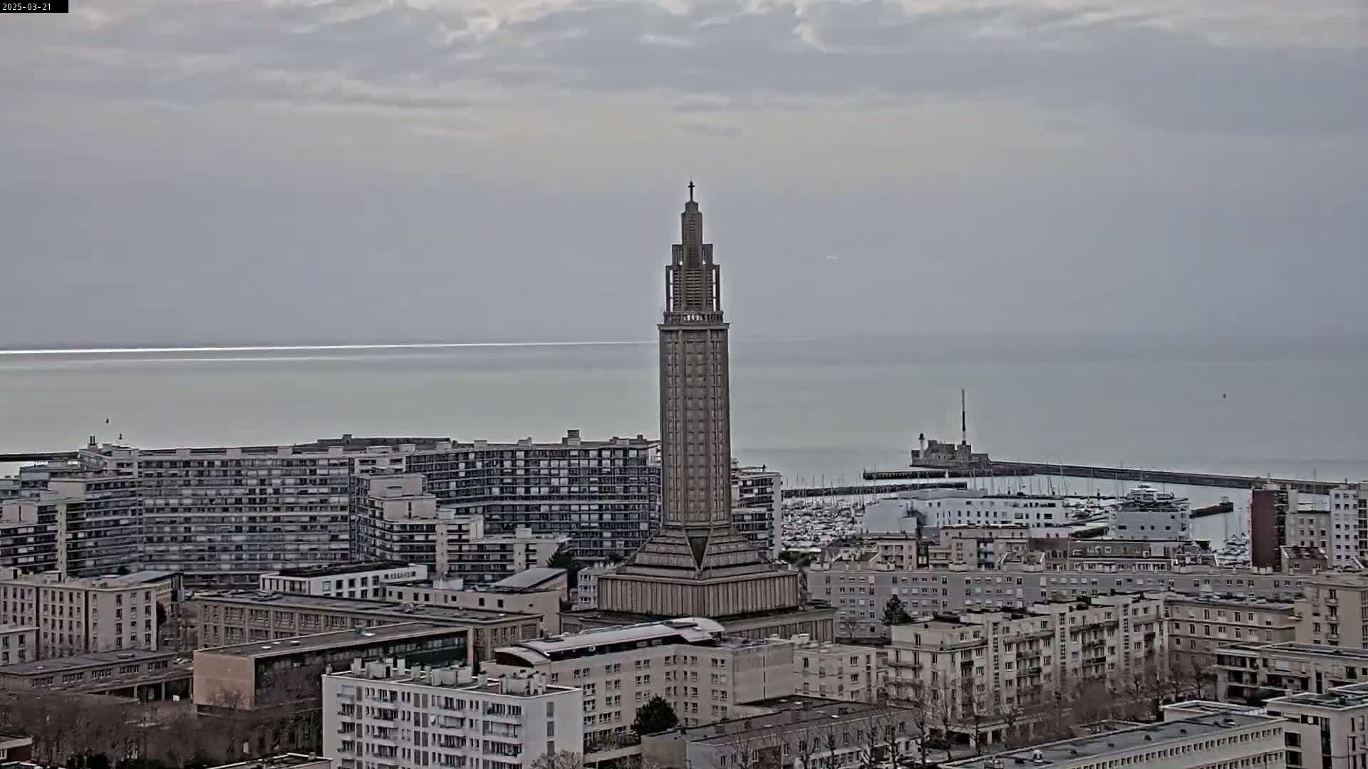 A tall, slender tower stands amidst a city overlooking a calm, gray sea under an overcast sky.