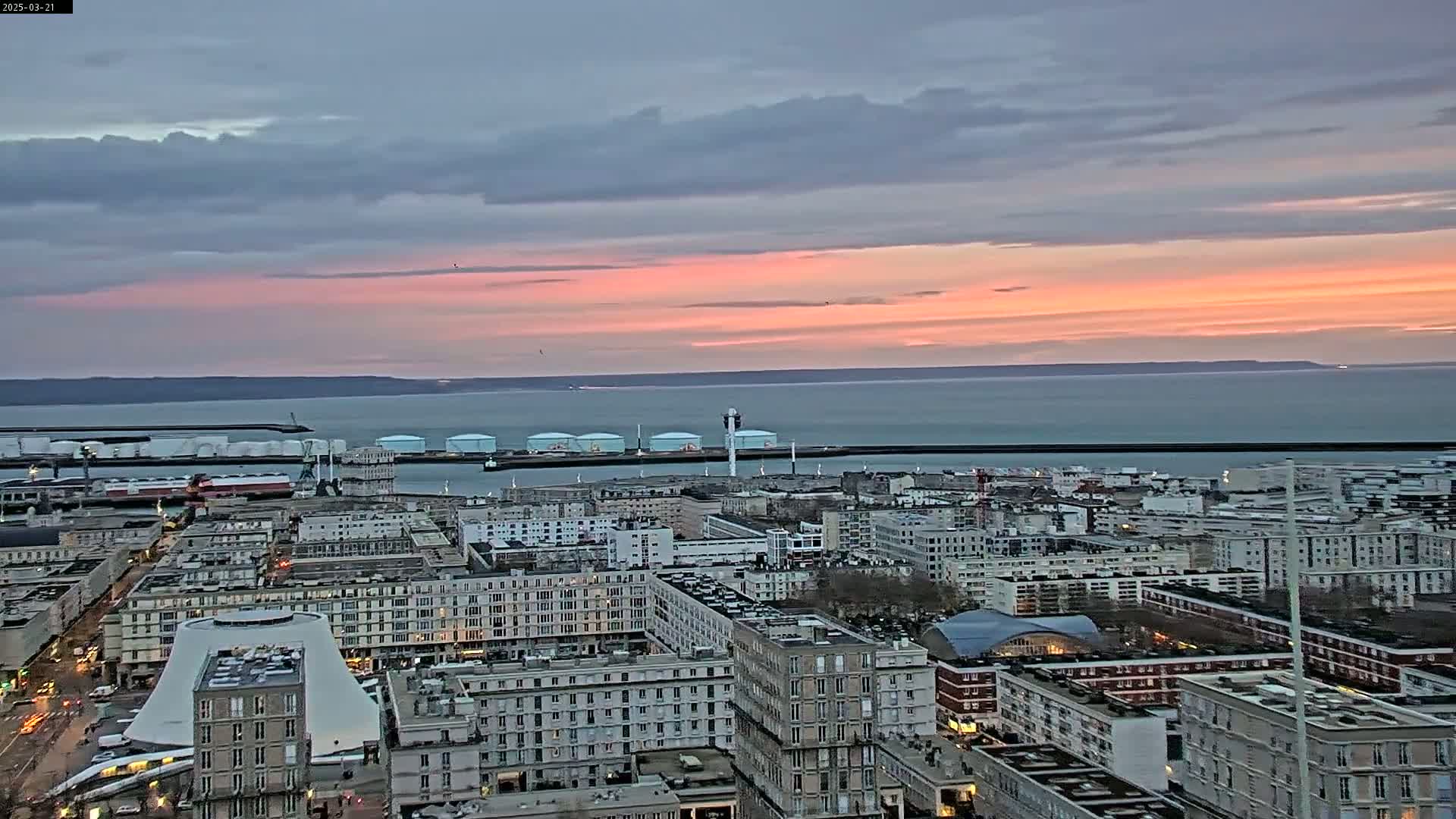 A coastal city at sunset, under a mostly cloudy sky, overlooks a calm body of water with a harbor containing industrial structures.