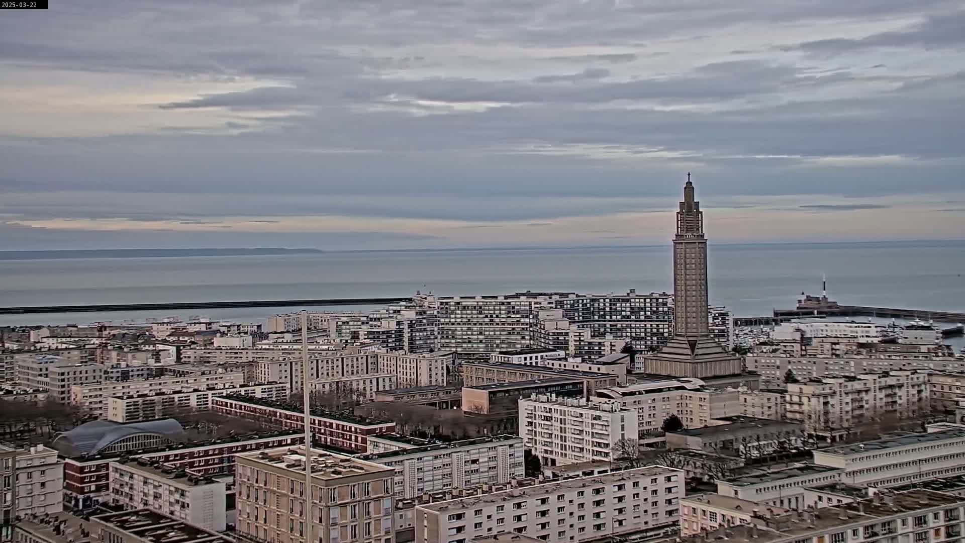 An aerial view shows a city with a tall, slender tower near a body of water under a cloudy sky.
