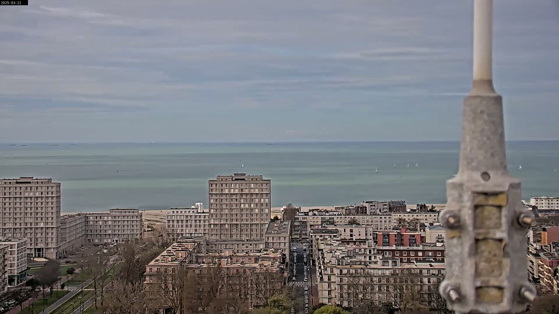 A coastal city with multi-story buildings stretches toward a calm, light-green sea under a cloudy sky.