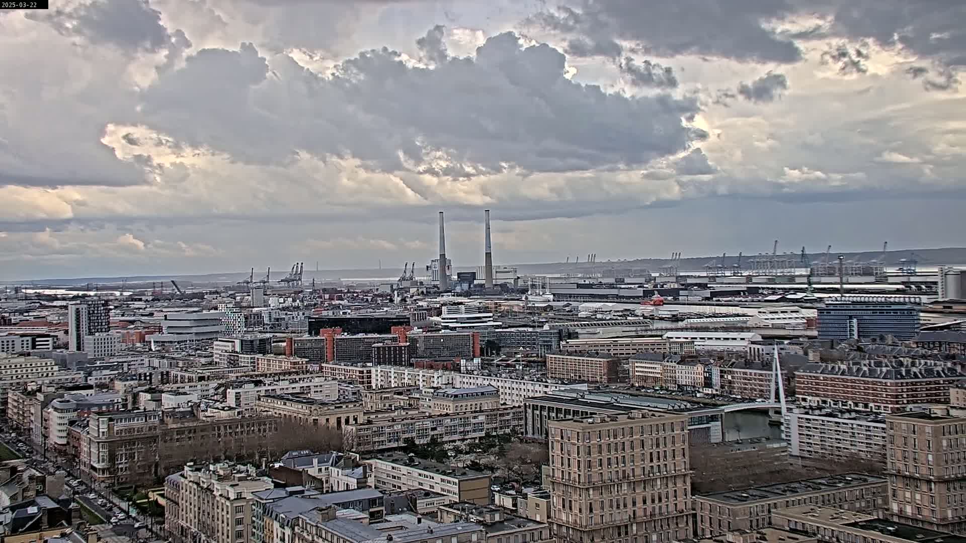 A sprawling cityscape under a cloudy sky, featuring a port with numerous cranes in the distance.