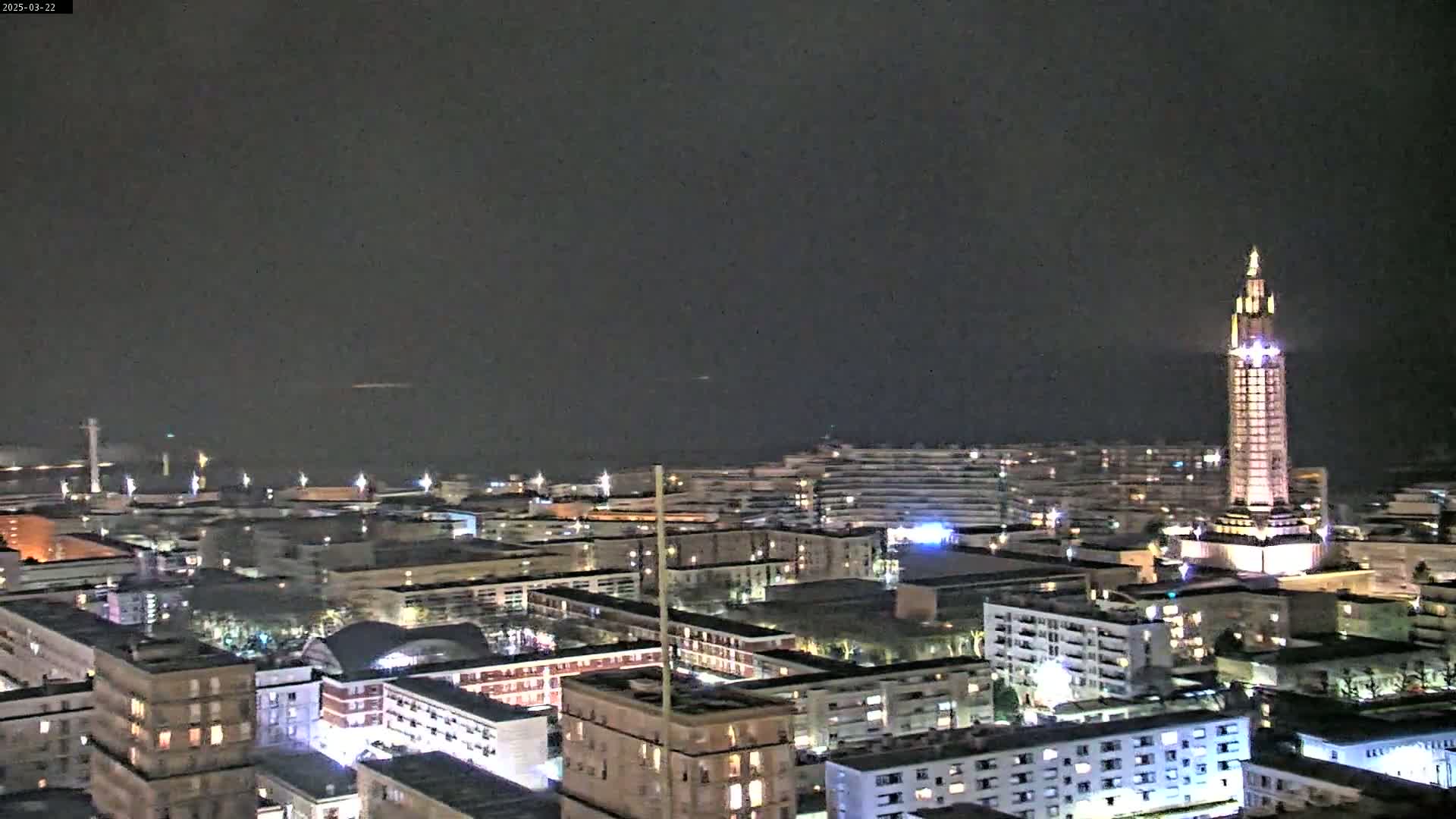 A nighttime cityscape with a tall illuminated tower is visible, under a dark, overcast sky.
