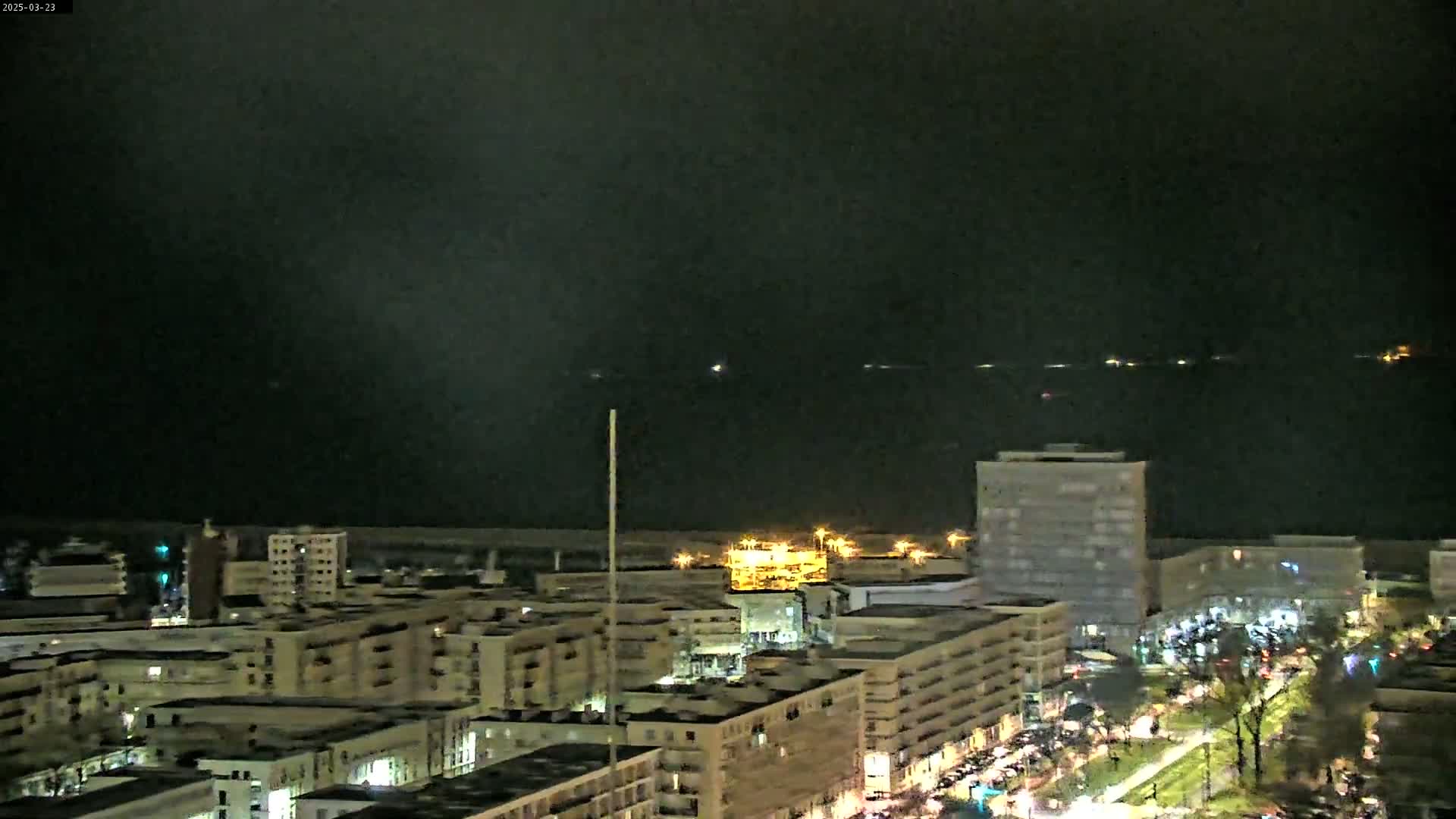 A nighttime view of a city near a dark body of water shows many lit buildings and roads under a cloudy sky.