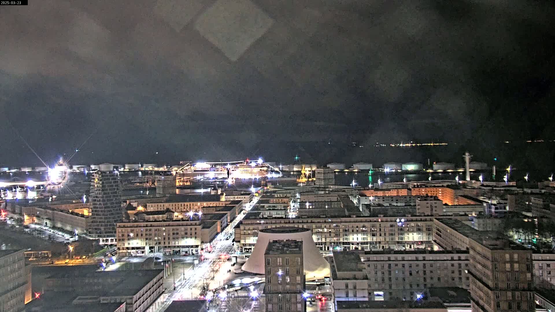 A nighttime aerial view of a city harbor, under a dark, cloudy sky, shows a large cruise ship docked near brightly lit buildings and industrial structures.