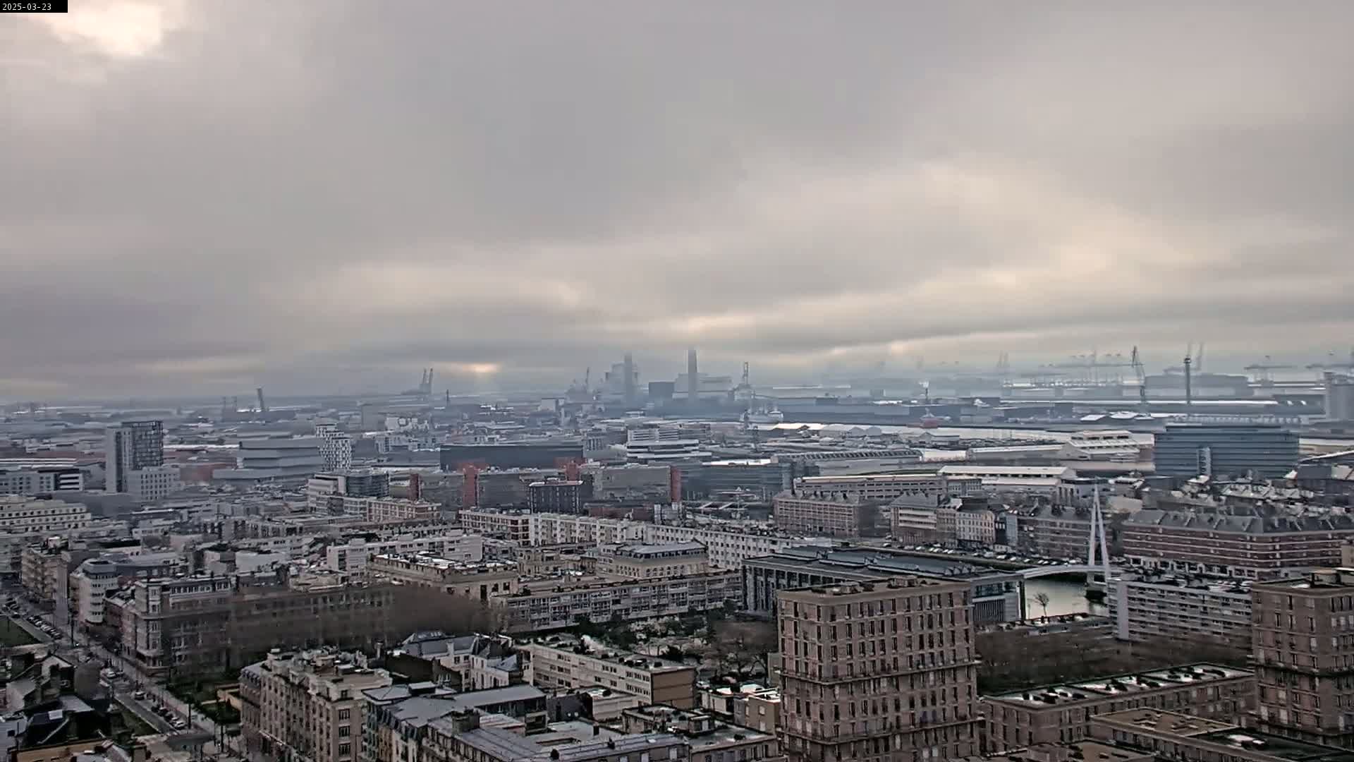 A hazy, overcast day reveals an expansive cityscape, featuring a dense collection of buildings that extend to an industrial port area in the distance.
