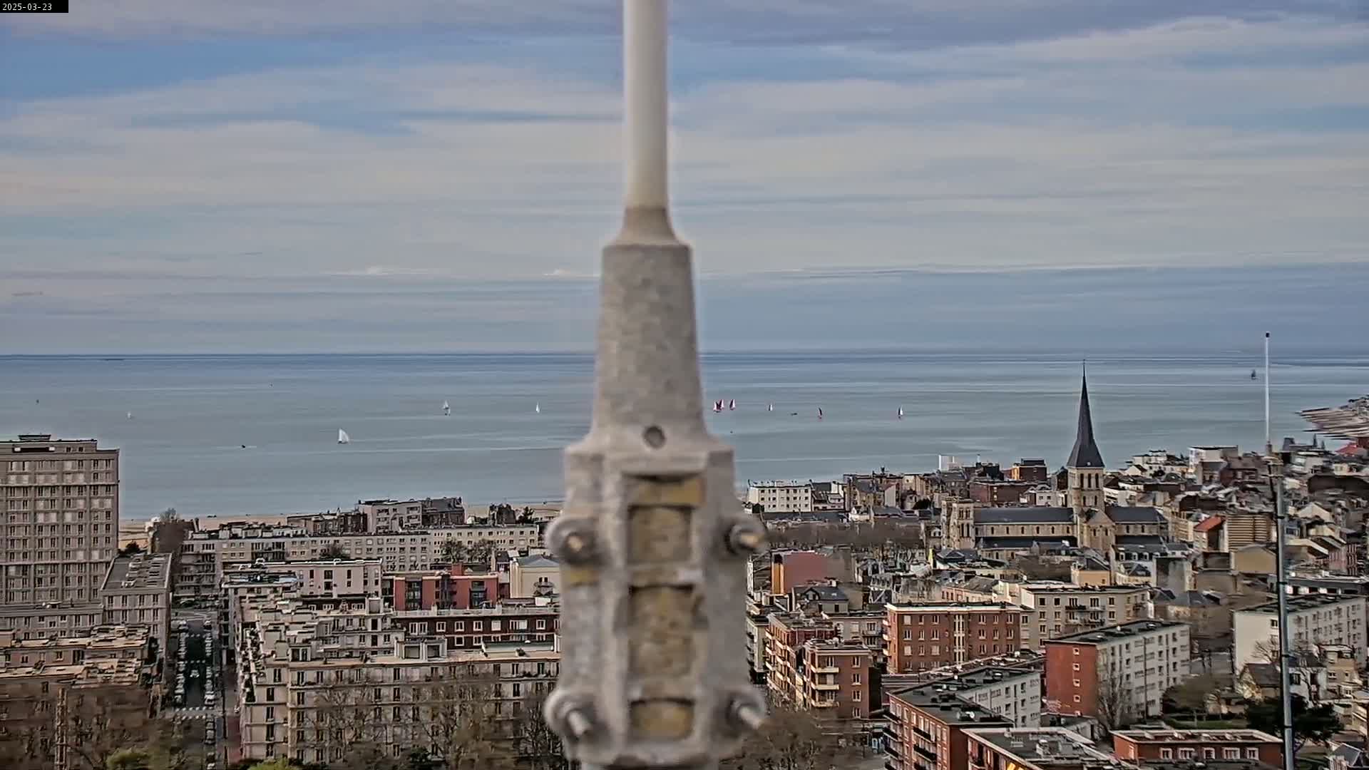 A gray metal pole stands in the foreground, overlooking a coastal city with a calm sea and several sailboats in the distance under a partly cloudy sky.