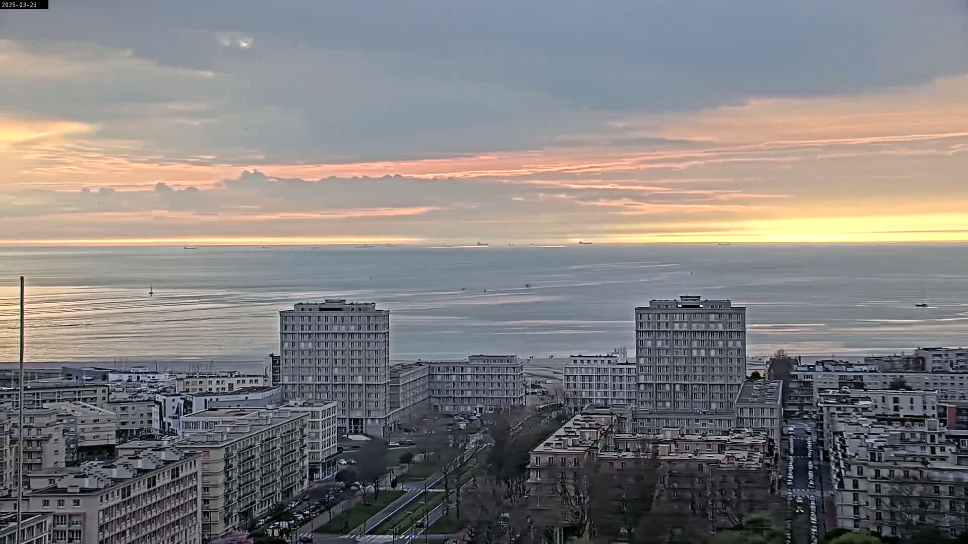 A city skyline with several high-rise buildings overlooks a calm ocean under a pale sunrise with thin clouds.