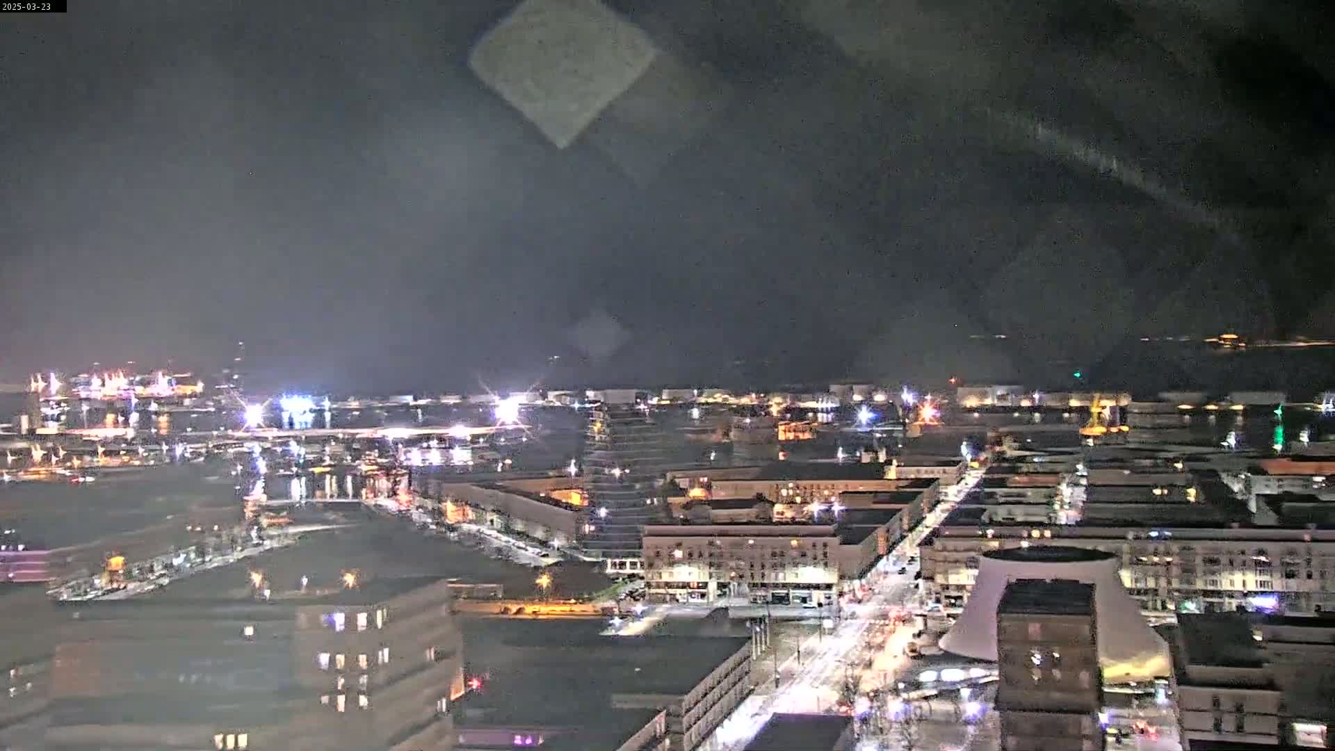 A nighttime, high-angle view of a city harbor and streets, with many lights illuminating buildings and roadways under a dark, cloudy sky.