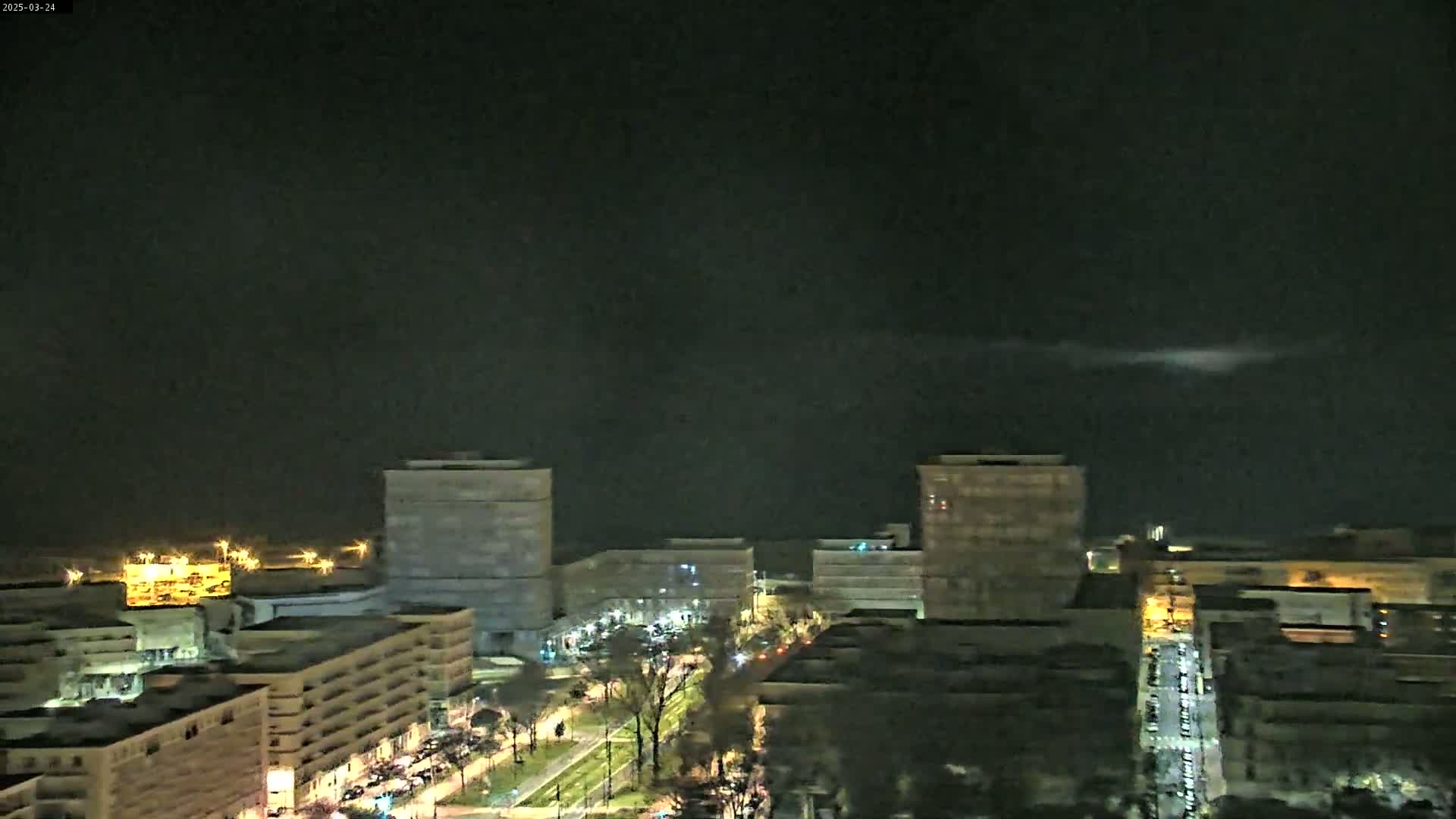 A nighttime, high-angle view shows a city with several multi-story buildings, illuminated streets, and a dark sky.
