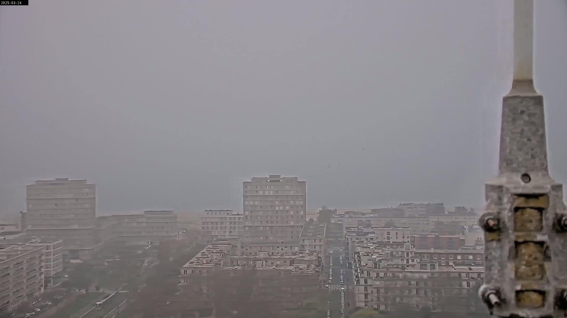 A hazy, overcast view shows a city with several multi-story buildings, a street running through the center, and sparse, low-lying vegetation.