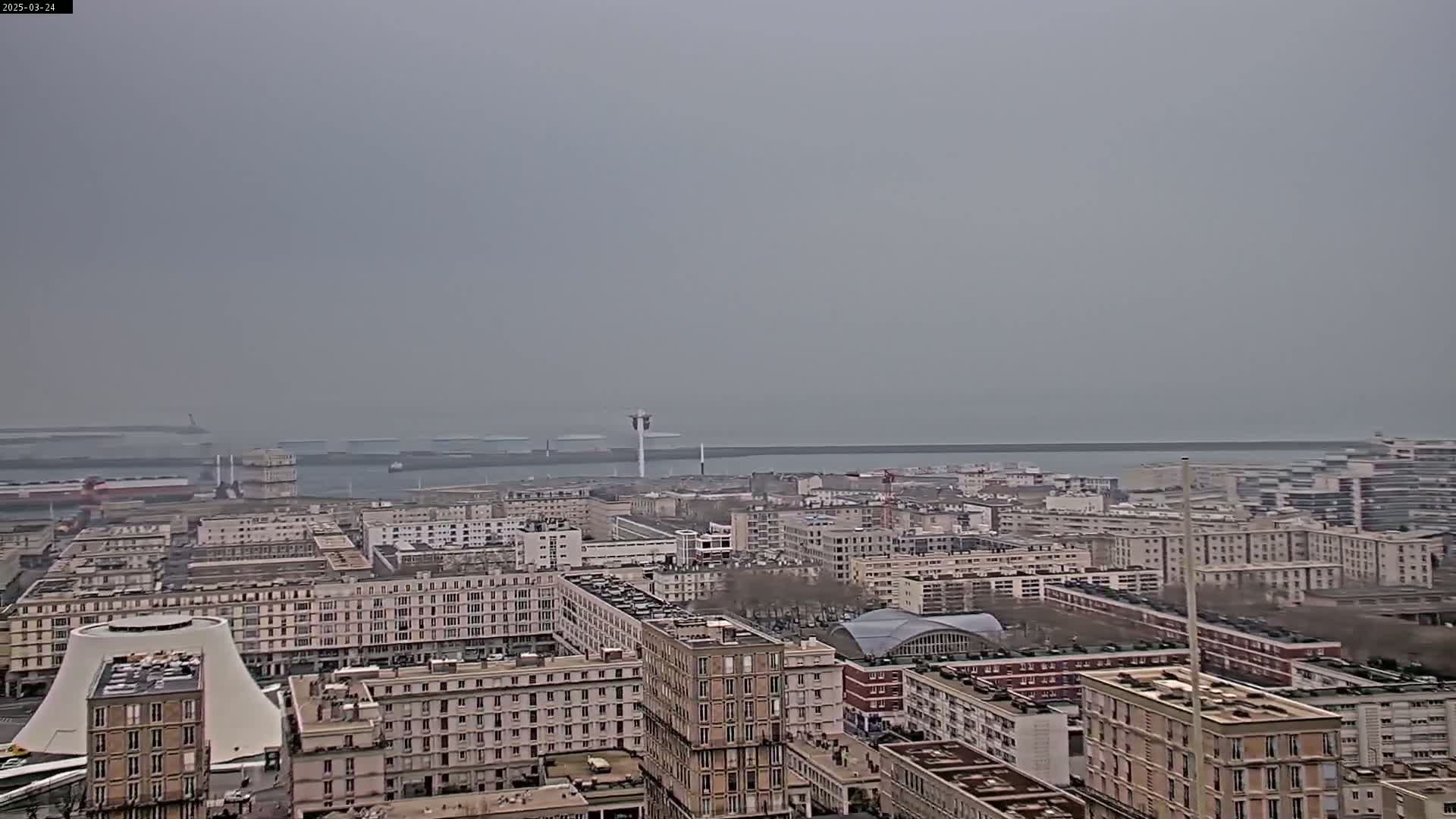 A hazy, overcast day reveals a cityscape overlooking a harbor with numerous ships and buildings.