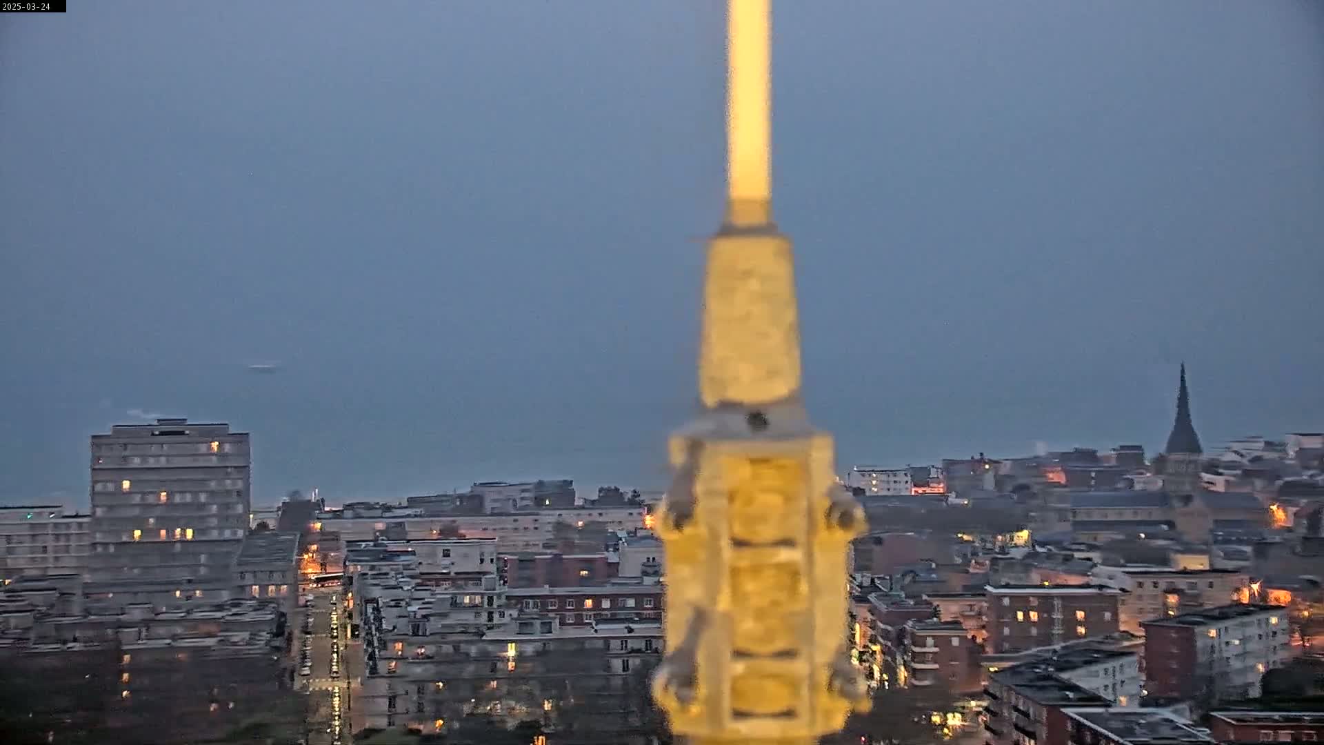 A blurry view of a city at dusk under an overcast sky, seen from a high vantage point through a tall, slender object in the foreground.