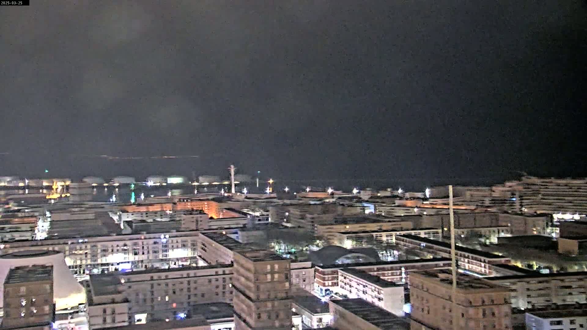 A nighttime aerial view of a city harbor with numerous buildings illuminated, under a dark, overcast sky.