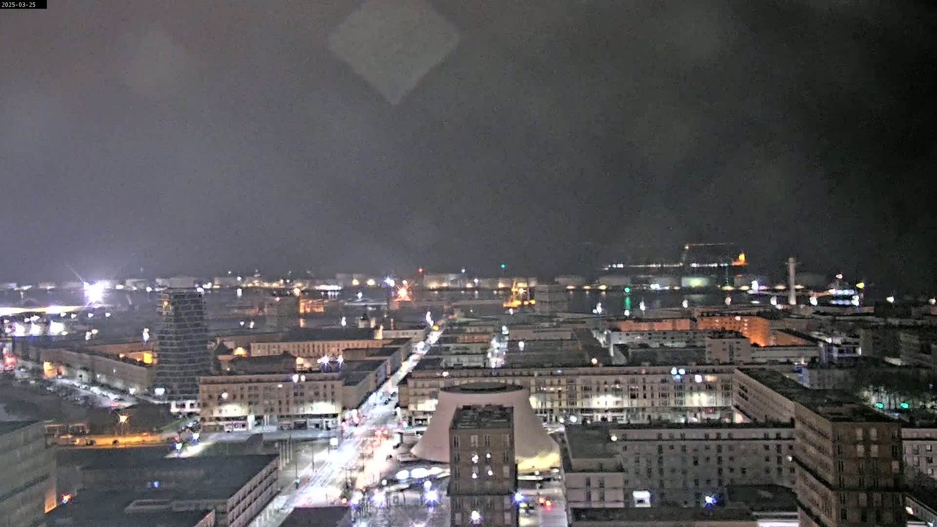 A nighttime, hazy view of a city with illuminated buildings and streets extending toward a harbor with lights visible in the distance.
