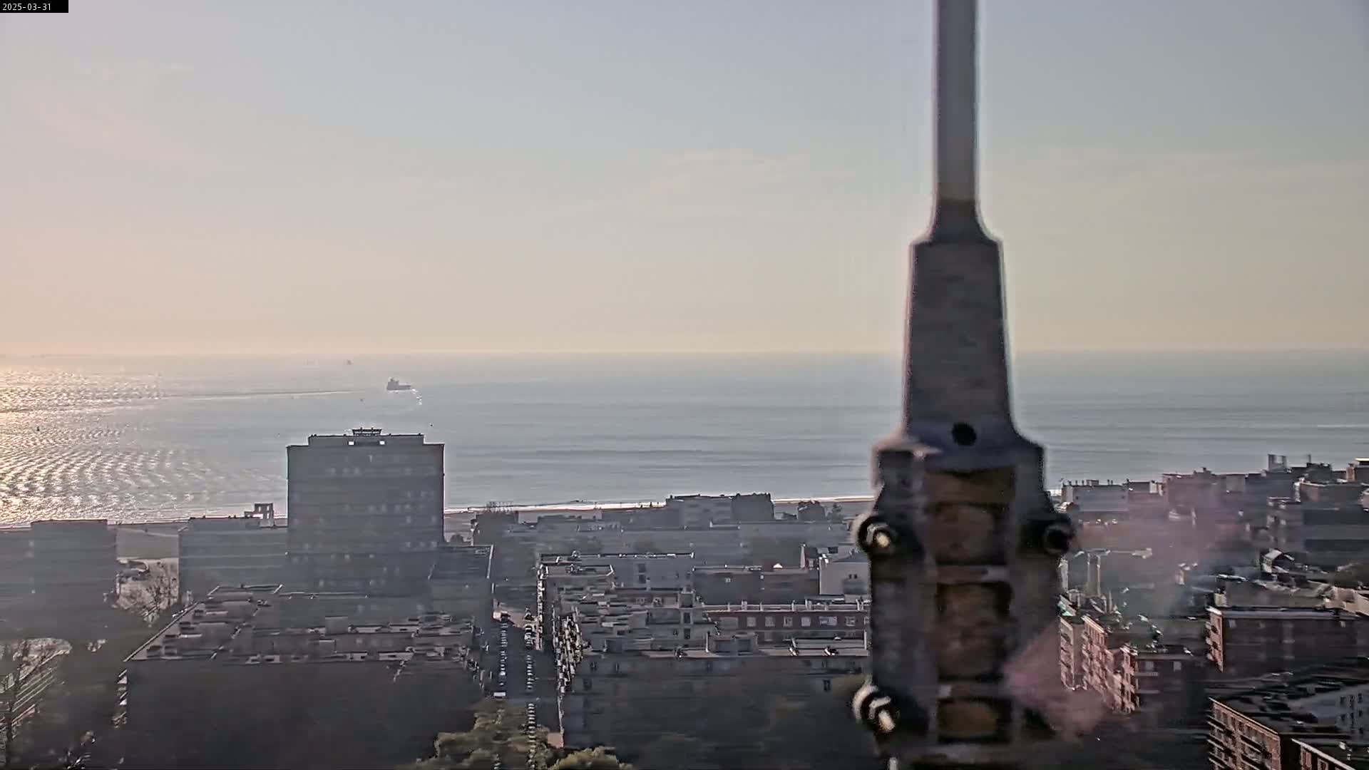 A hazy, sunny day overlooks a city near a calm ocean with a few ships visible in the distance, partially obscured by a tall structure in the foreground.