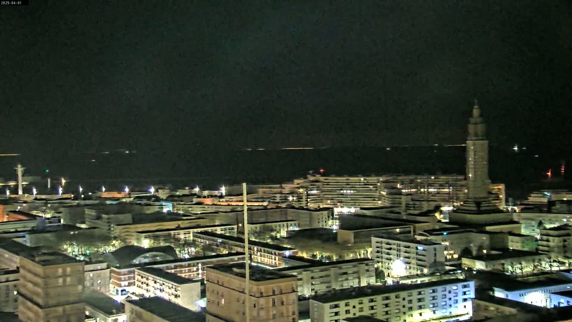 A nighttime aerial view of a city near a calm body of water, showing numerous buildings with lights on.