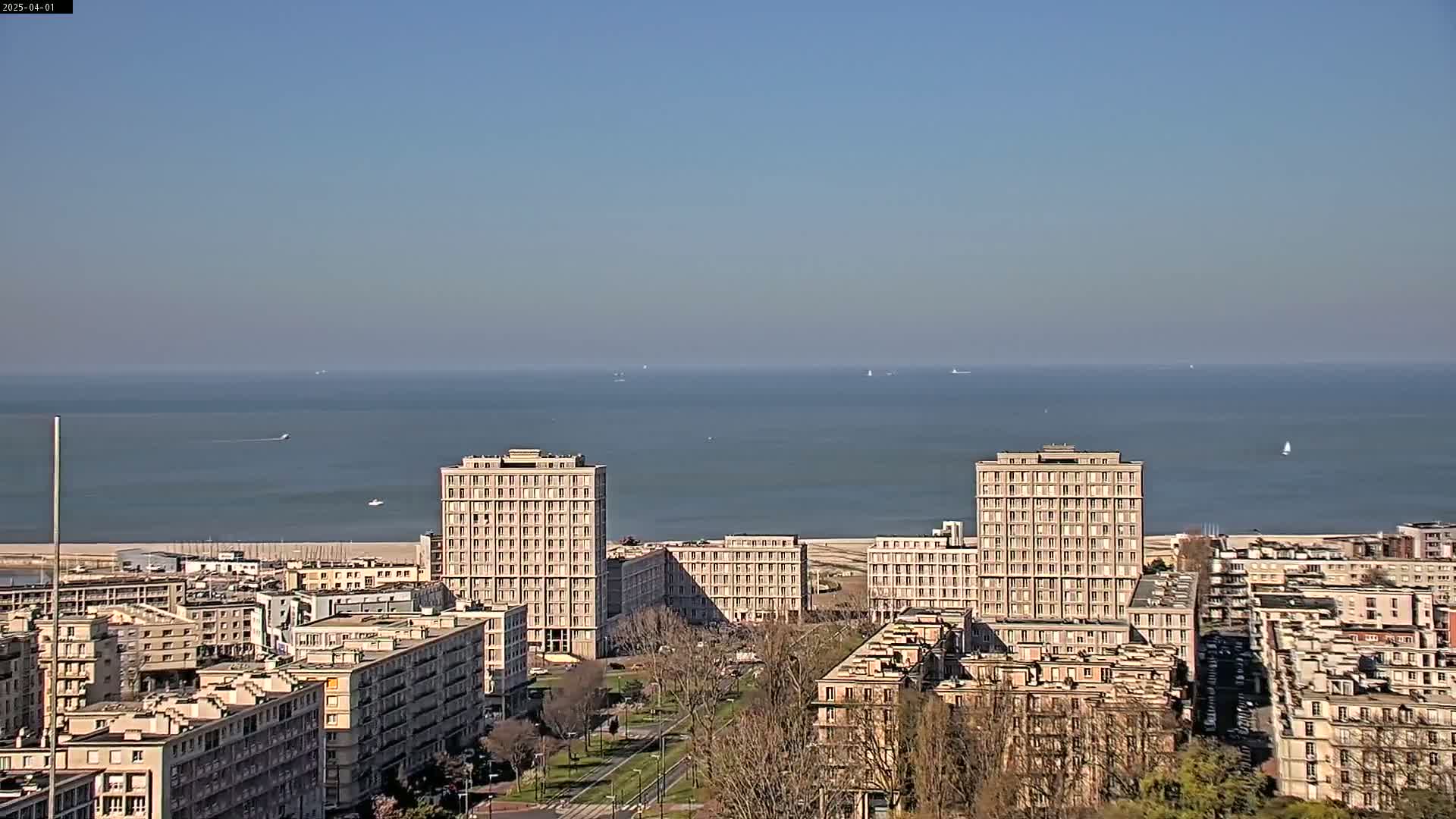 A cityscape with several high-rise buildings is situated near a calm ocean under a clear, blue sky.