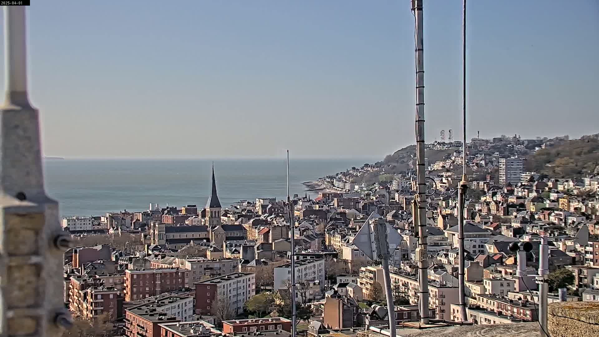 A coastal city with many buildings and a church steeple is visible on a clear, sunny day, with a body of water in the background.